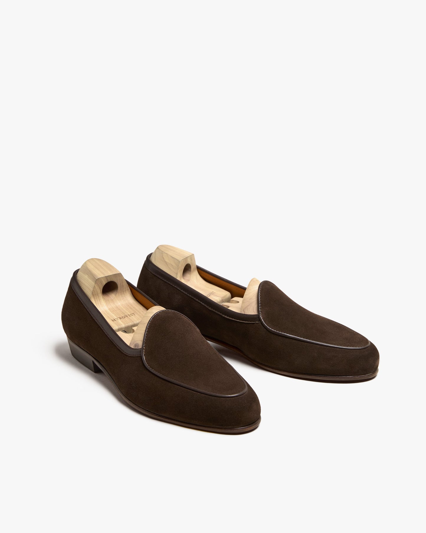 Ängby in dark brown suede displayed at a three-quarter angle, featuring a Belgian-inspired loafer silhouette, soft suede finish, apron piping, and wooden shoe trees inside each shoe.