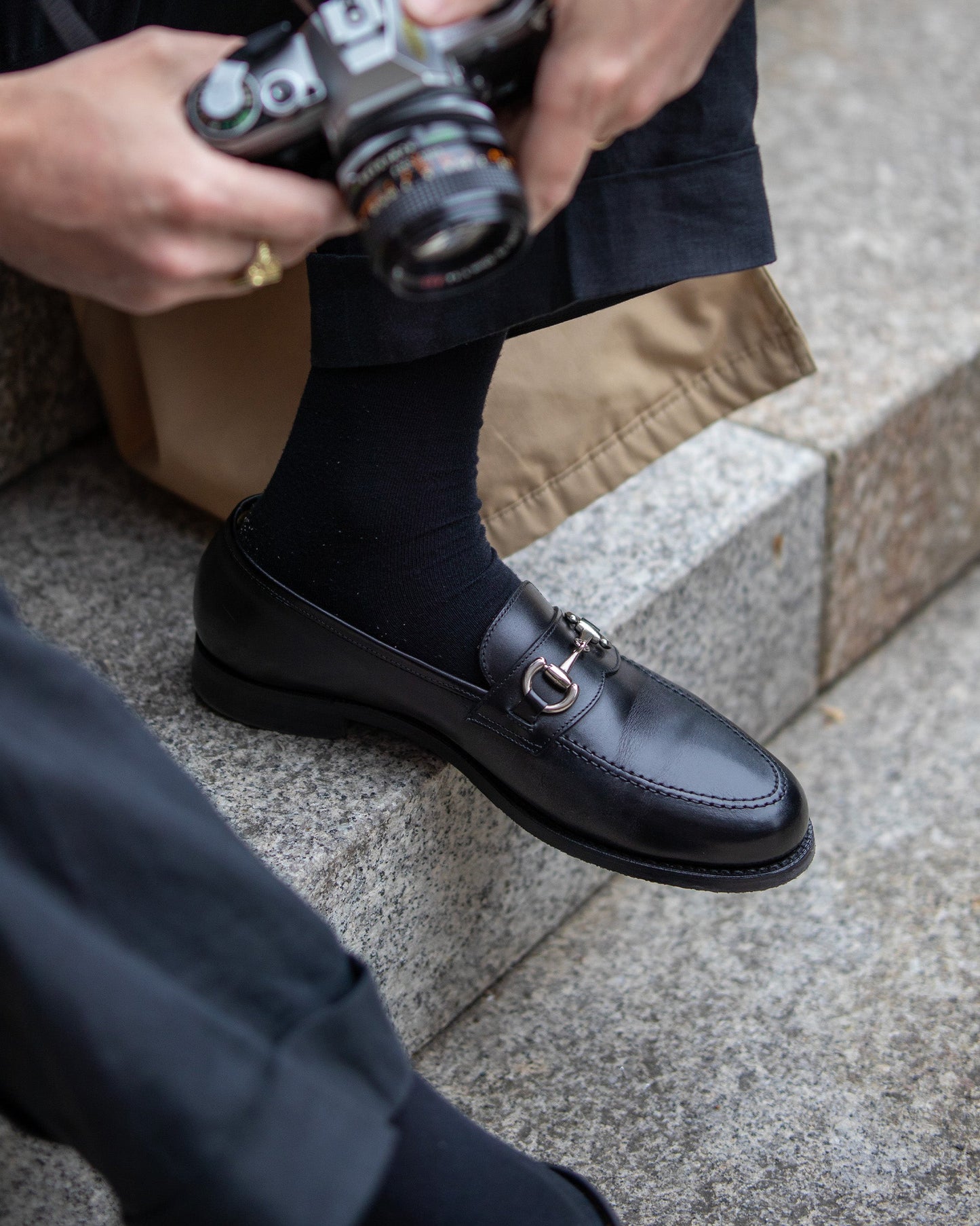 Karlberg in black calf leather worn with dark trousers and black socks, featuring a polished finish and silver-toned horsebit detail, styled for a casual city setting with a camera in hand.