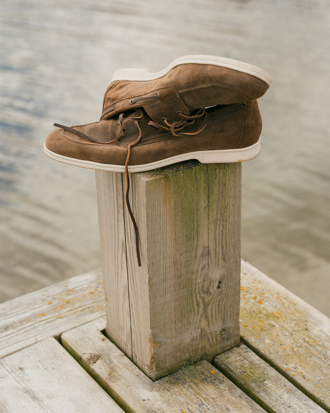 Sandhamn in chestnut waxed suede shows a pair of brown suede loafers with white soles and understated lacing, casually stacked on a wooden dock post beside the water, highlighting their relaxed, maritime-inspired style.