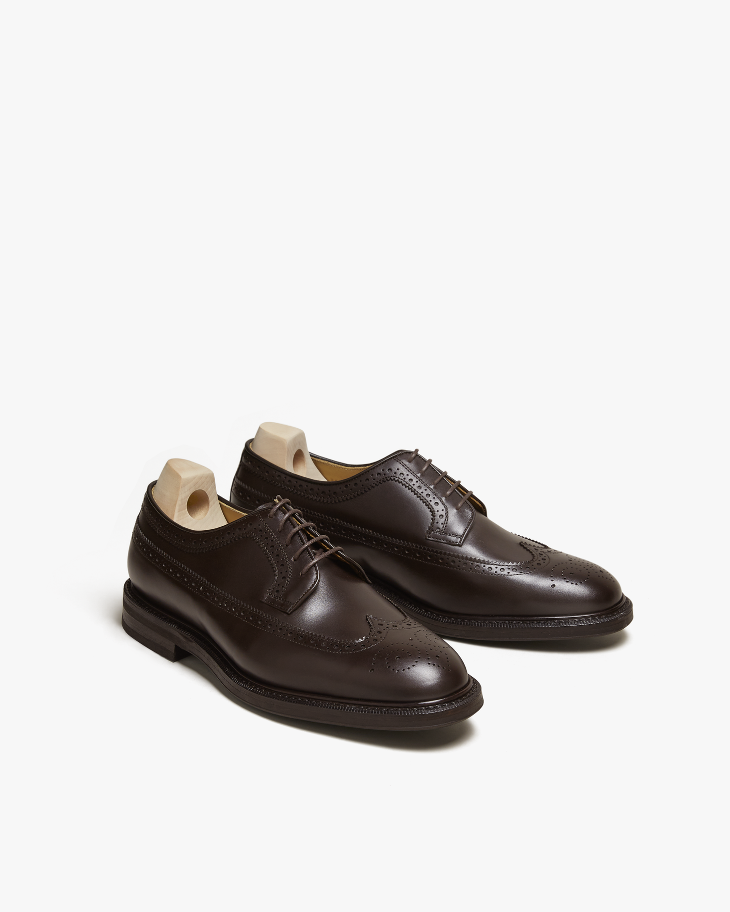 Orust II in dark brown calf, a pair of men's derby shoes with wingtip brogue detailing and open lacing, displayed at an angle with shoe trees to emphasize the polished finish and elegant silhouette.