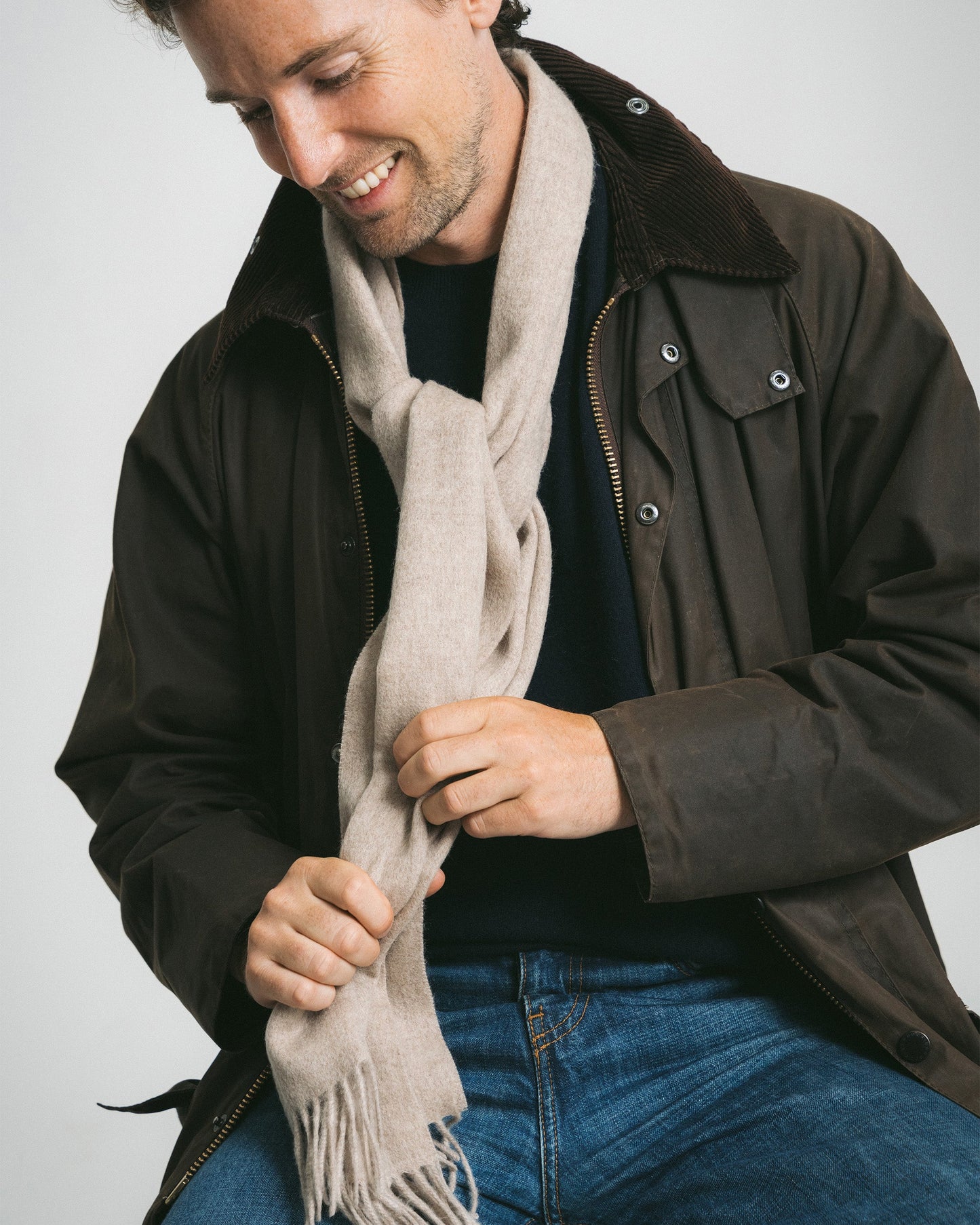 A smiling man adjusts a beige wool scarf worn with a dark brown jacket, sweater, and blue jeans.