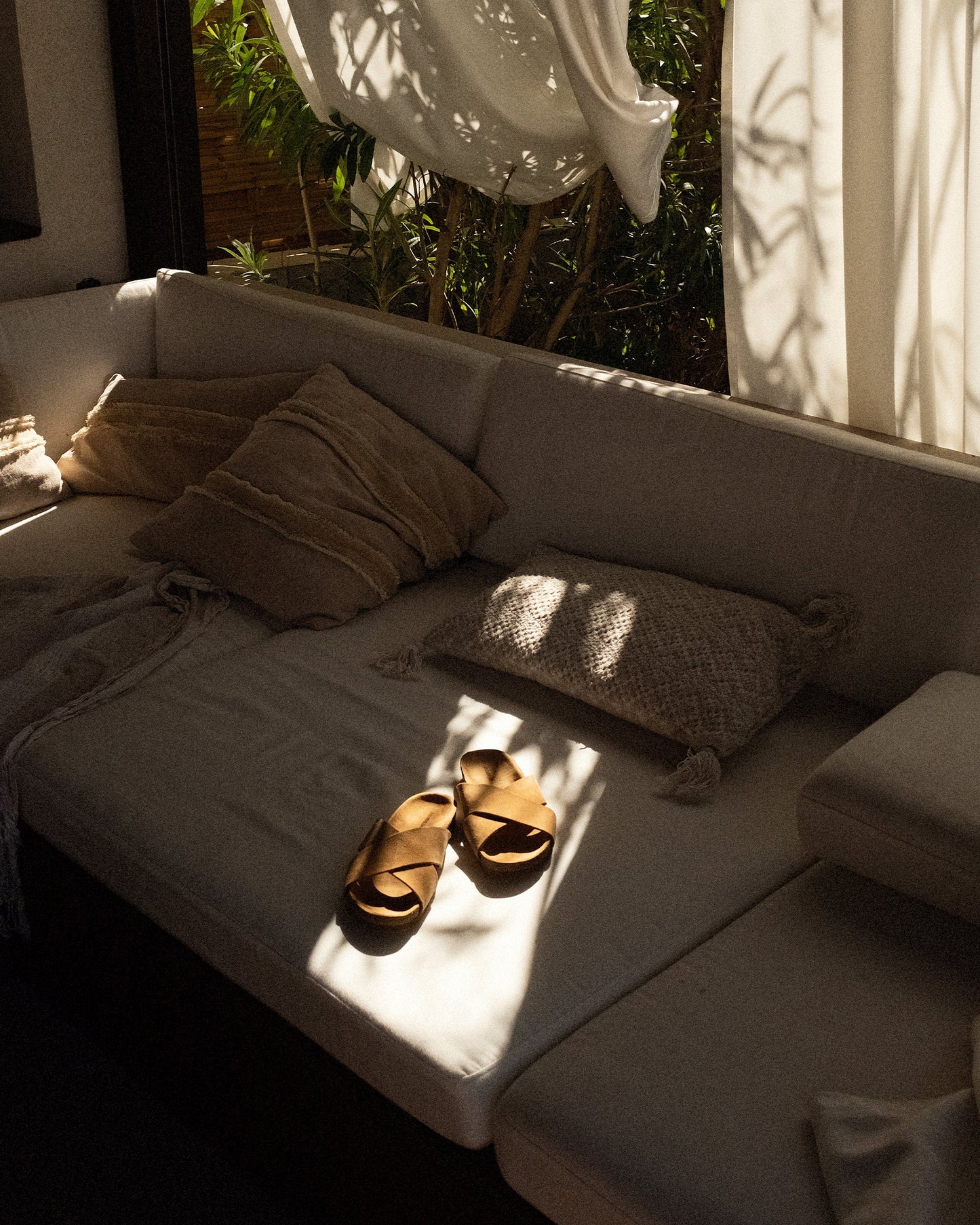 Solvik in sand suede, with wide cross-over suede straps and a cork sole, rests on a sunlit outdoor sofa surrounded by neutral pillows and soft natural light.