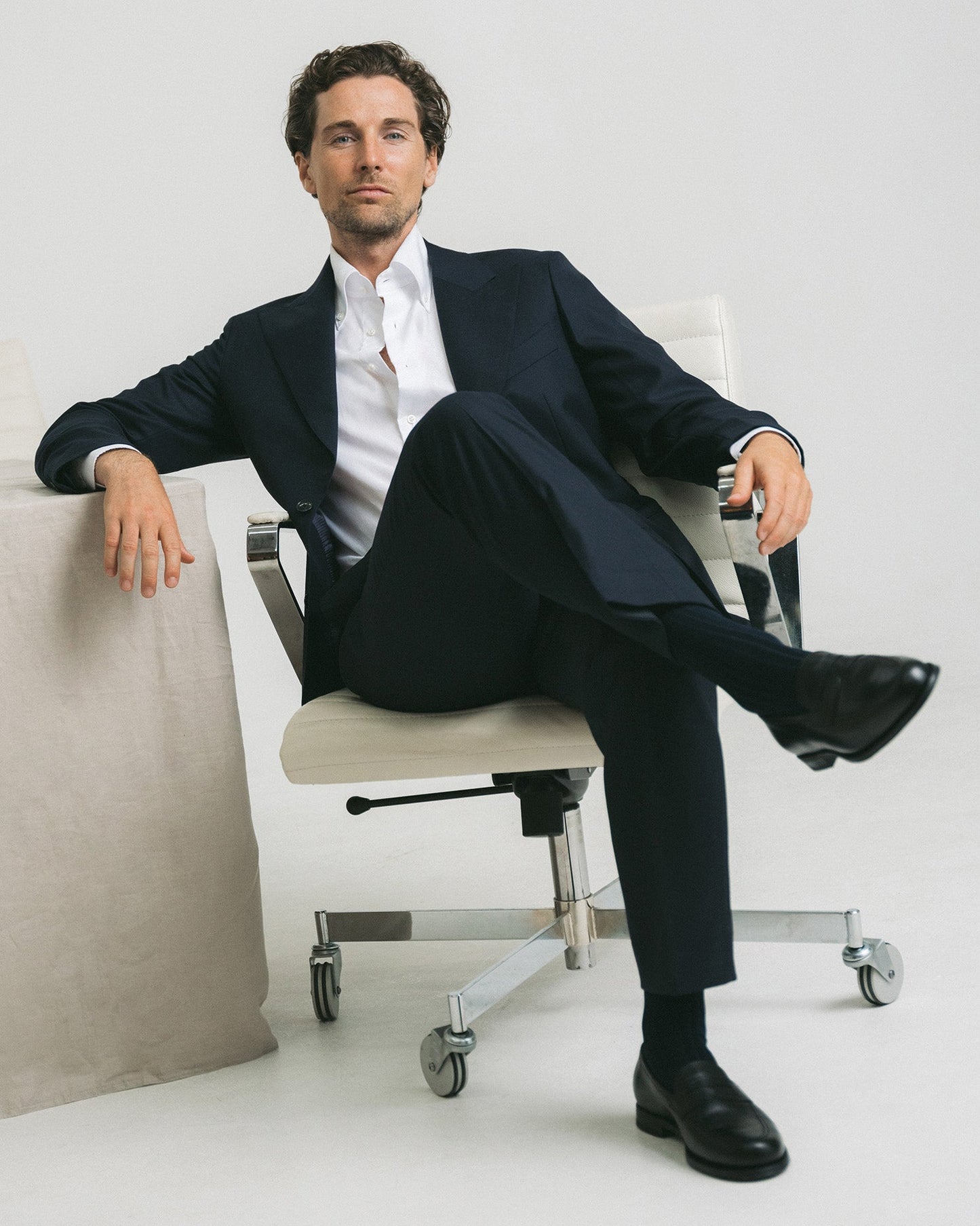 A man sits in a modern office chair wearing Stenhammar II in black calf, paired with a tailored navy suit and dark socks, highlighting the loafer’s polished finish and classic penny strap style for a refined business look.