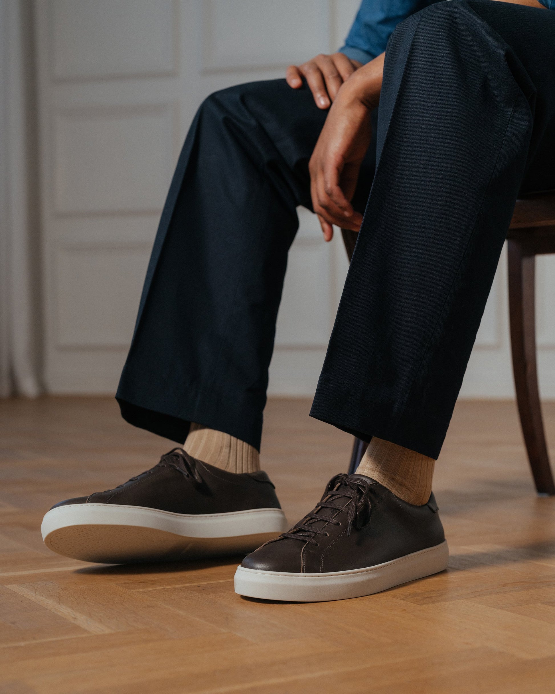 Oaxen in dark brown calf is worn with tailored dark trousers and beige socks, highlighting the refined brown leather and white rubber sole in a relaxed, indoor setting.