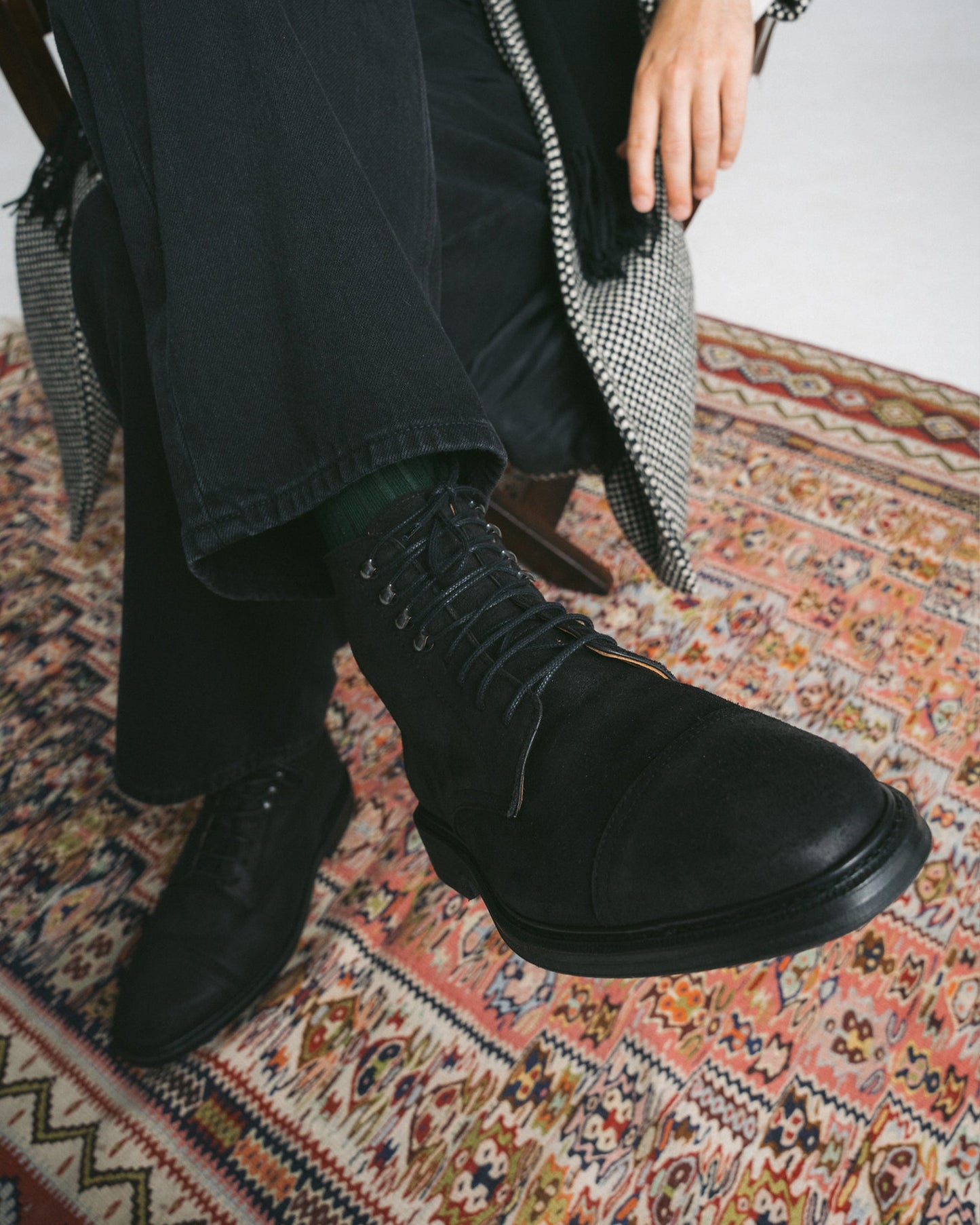 A man sits cross-legged on a patterned rug, wearing Abisko in black waxed suede styled with dark trousers and a checkered coat, highlighting the boot’s matte finish, cap toe design, and sturdy lace-up construction.