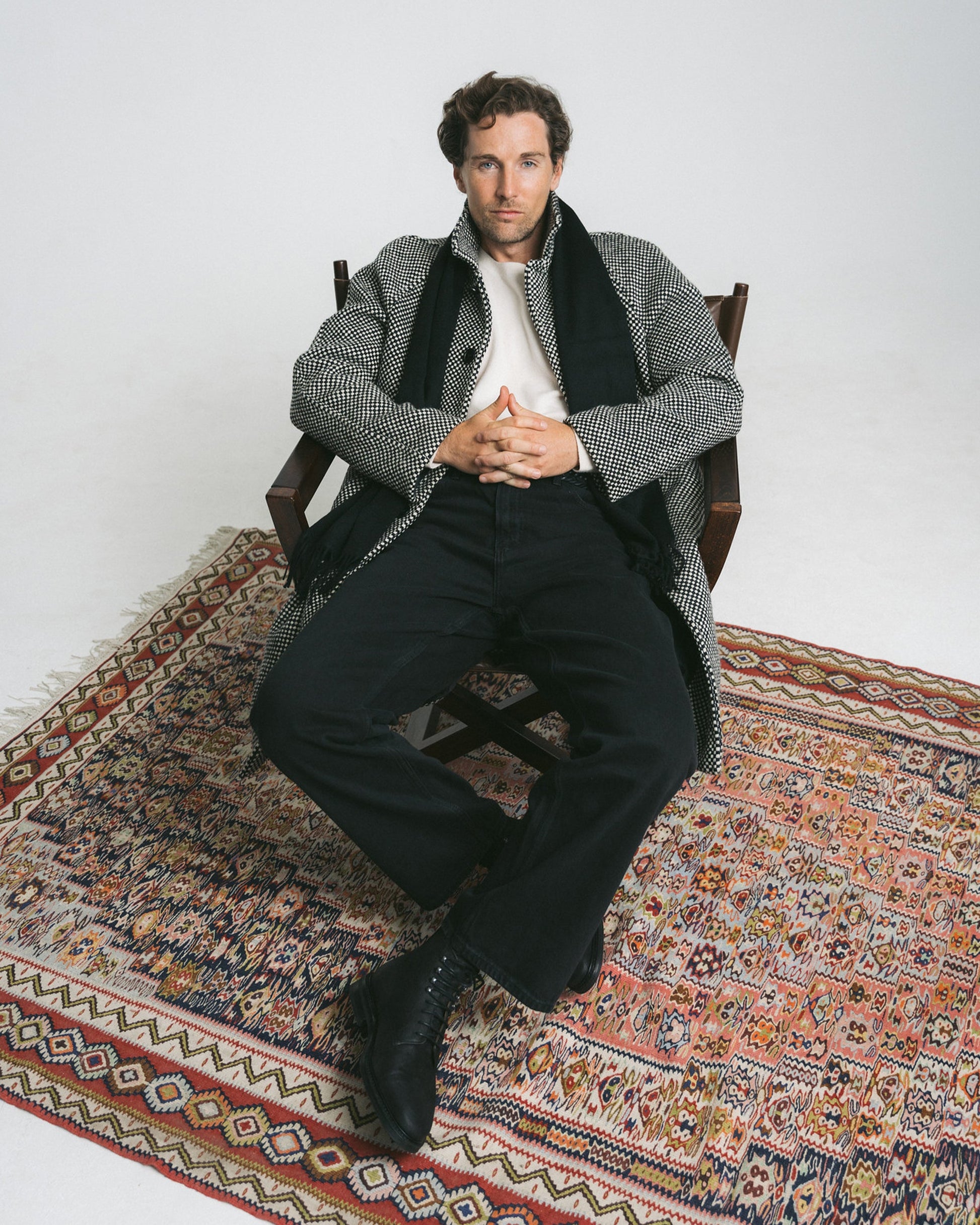 A man sits on a patterned rug wearing Abisko in black waxed suede, styled with black trousers, a white sweater, and a checkered overcoat, showcasing the boots’ matte finish and versatile winter look.