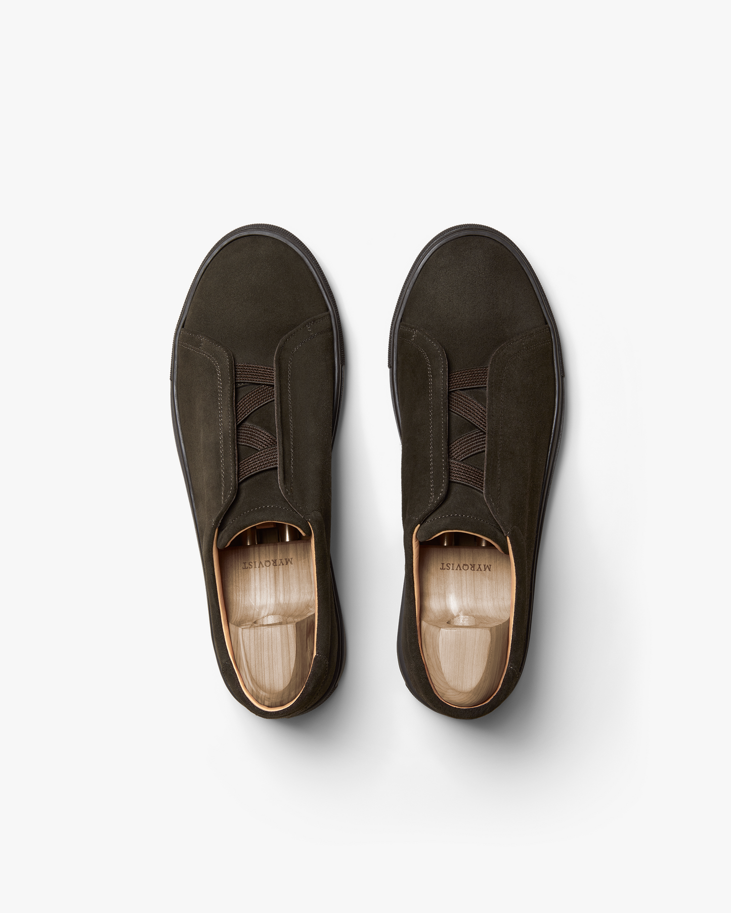 Pair of Alnö Monochrome II slip-on sneakers in dark brown suede shown from above, highlighting the seamless monochrome design, tonal stitching, and elastic instep bands with wooden shoe trees inside.