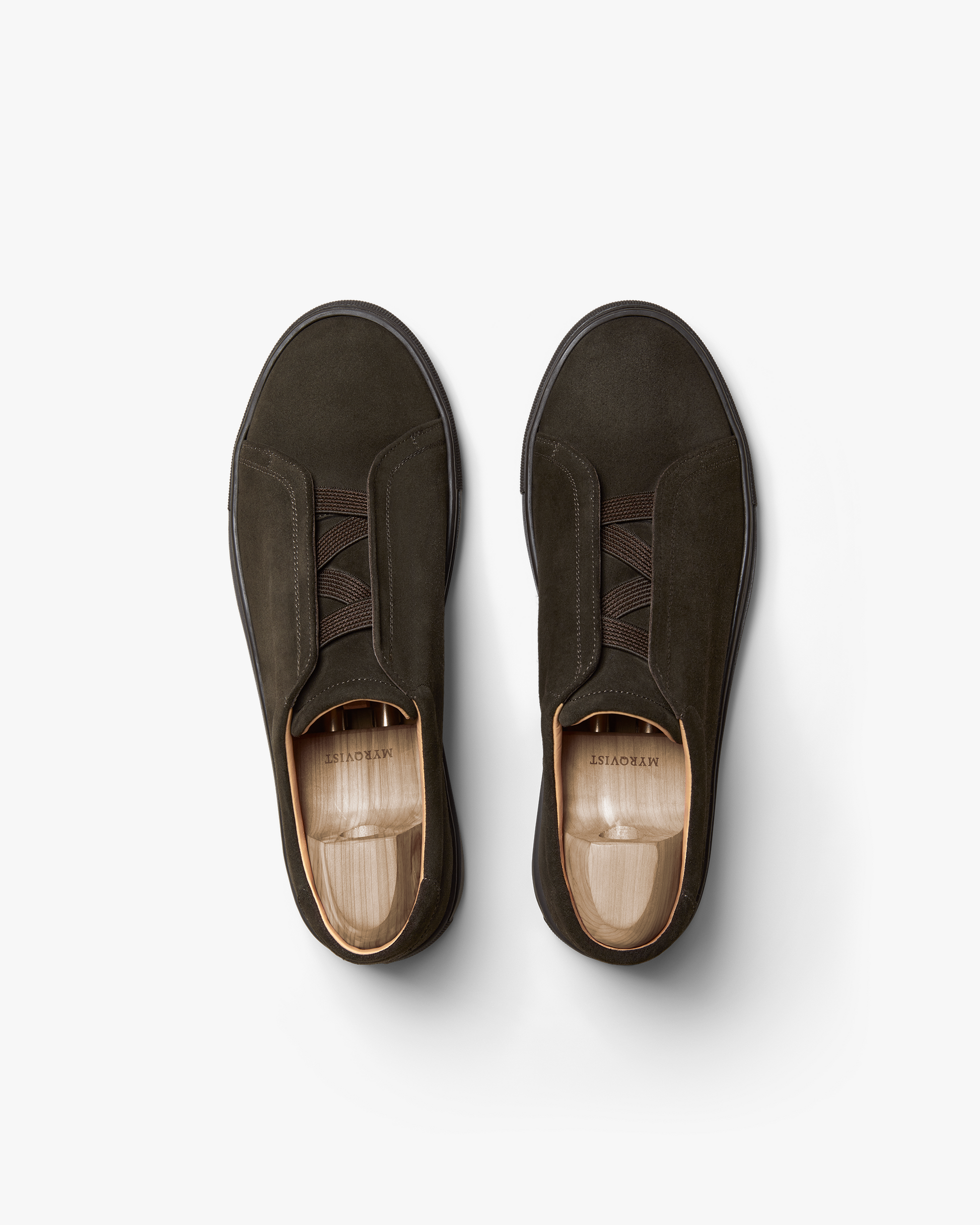 Pair of Alnö Monochrome II slip-on sneakers in dark brown suede shown from above, highlighting the seamless monochrome design, tonal stitching, and elastic instep bands with wooden shoe trees inside.
