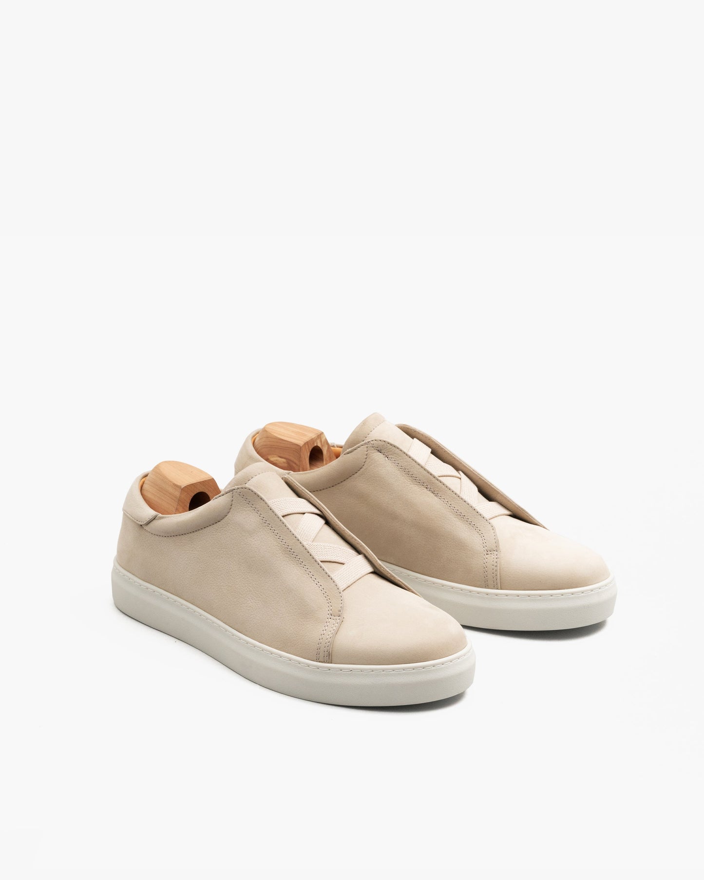 Pair of Alnö II in beige nubuck featuring a minimalist slip-on design with elastic crossover detail, softly textured uppers, and clean white cup soles, each displayed with a wooden shoe tree.
