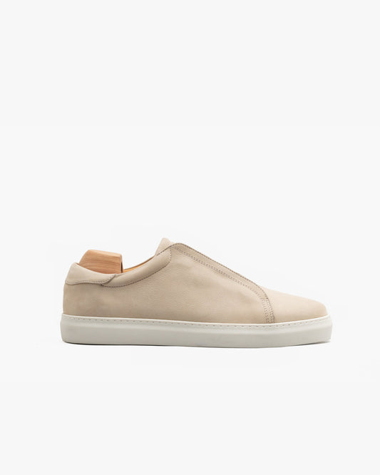 Minimalist low-top sneaker in beige nubuck with a seamless laceless design, subtle elastic crossover, and clean white cup sole, shown in profile with a wooden shoe tree inserted.