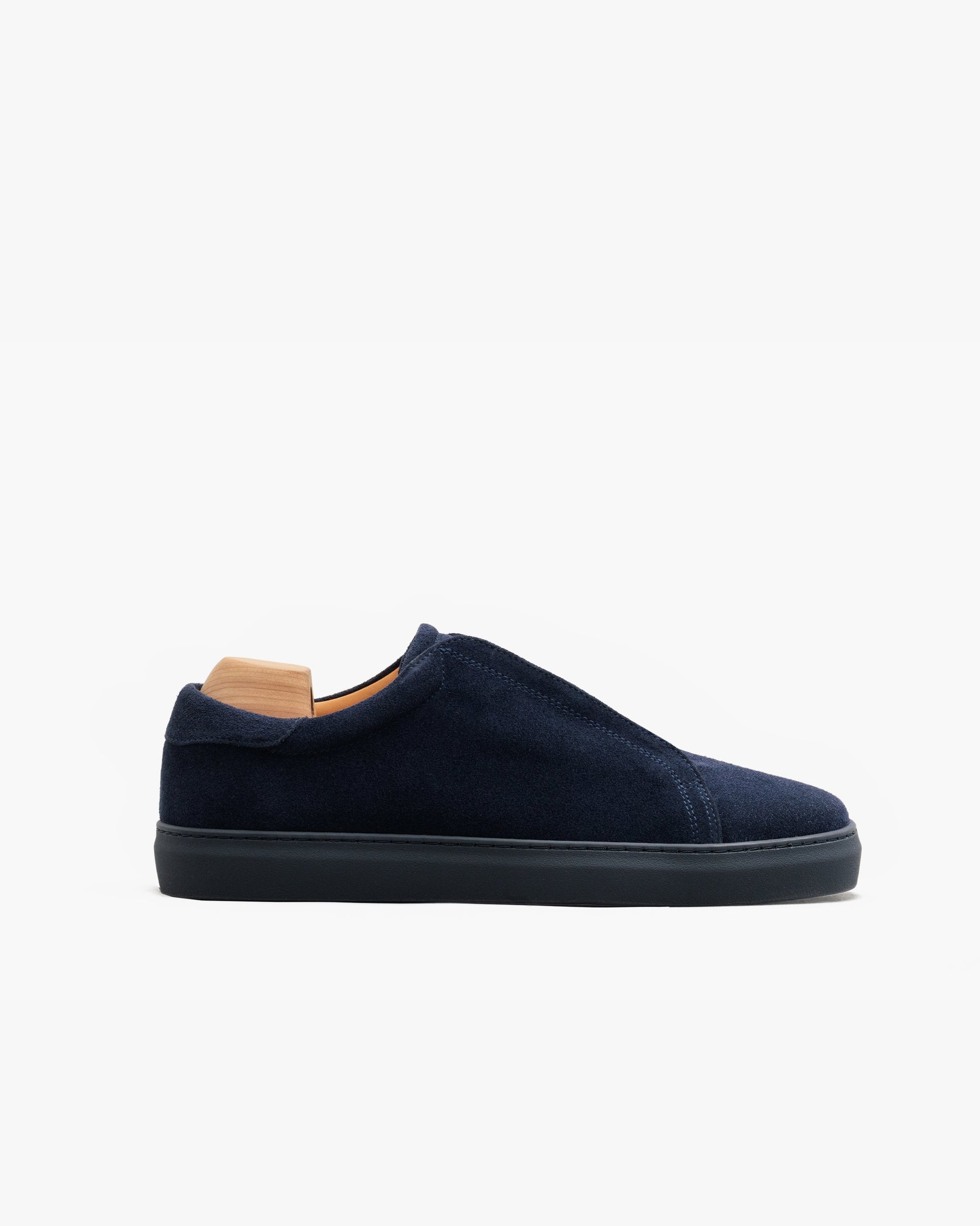 Low-top slip-on sneaker in midnight blue suede with a clean, seamless upper, tonal rubber sole, and a wooden shoe tree inserted for shape.