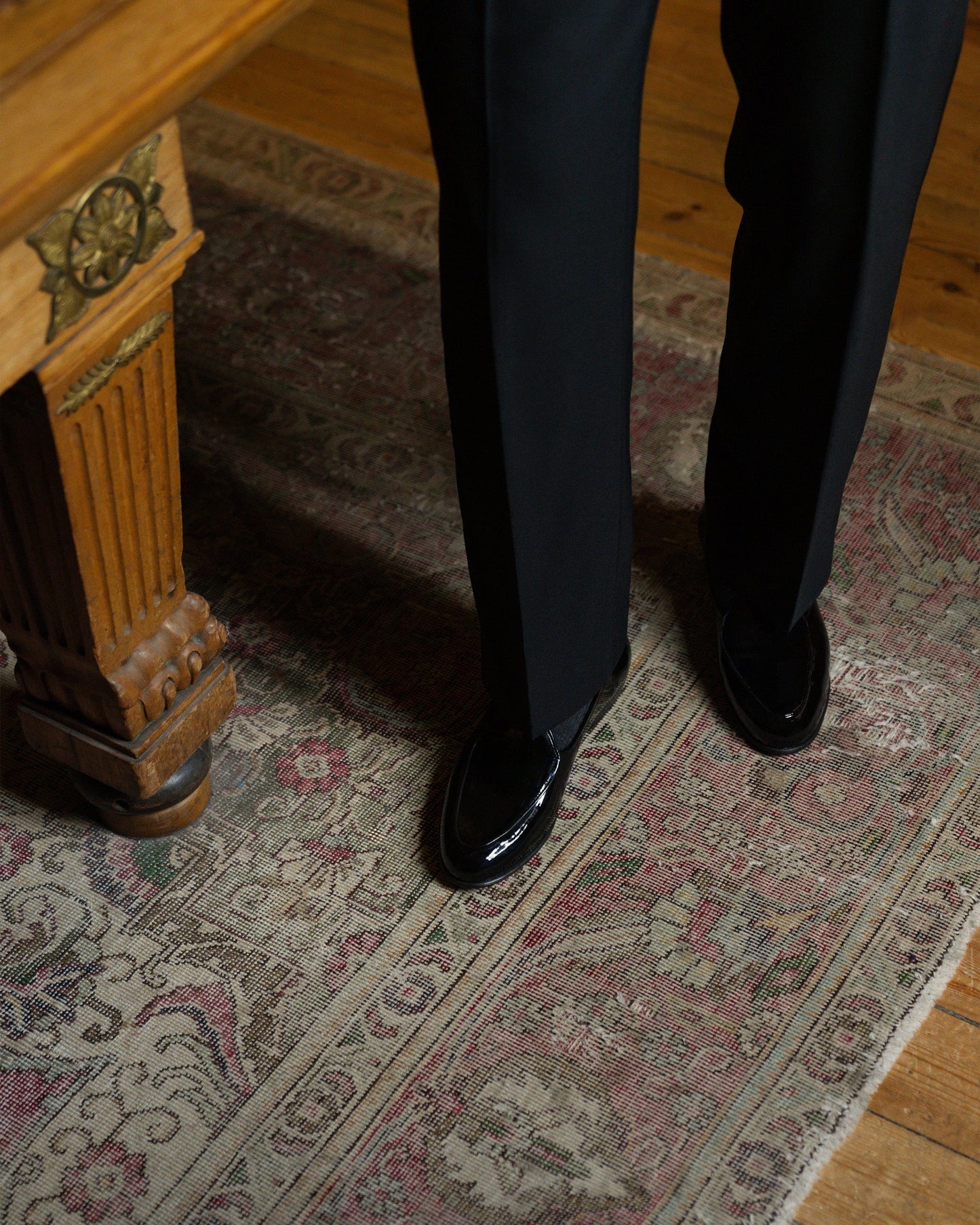 A man wearing Ängby in black patent leather stands on an ornate rug, pairing the high-shine Belgian loafers with tailored black trousers for an elegant formal look.