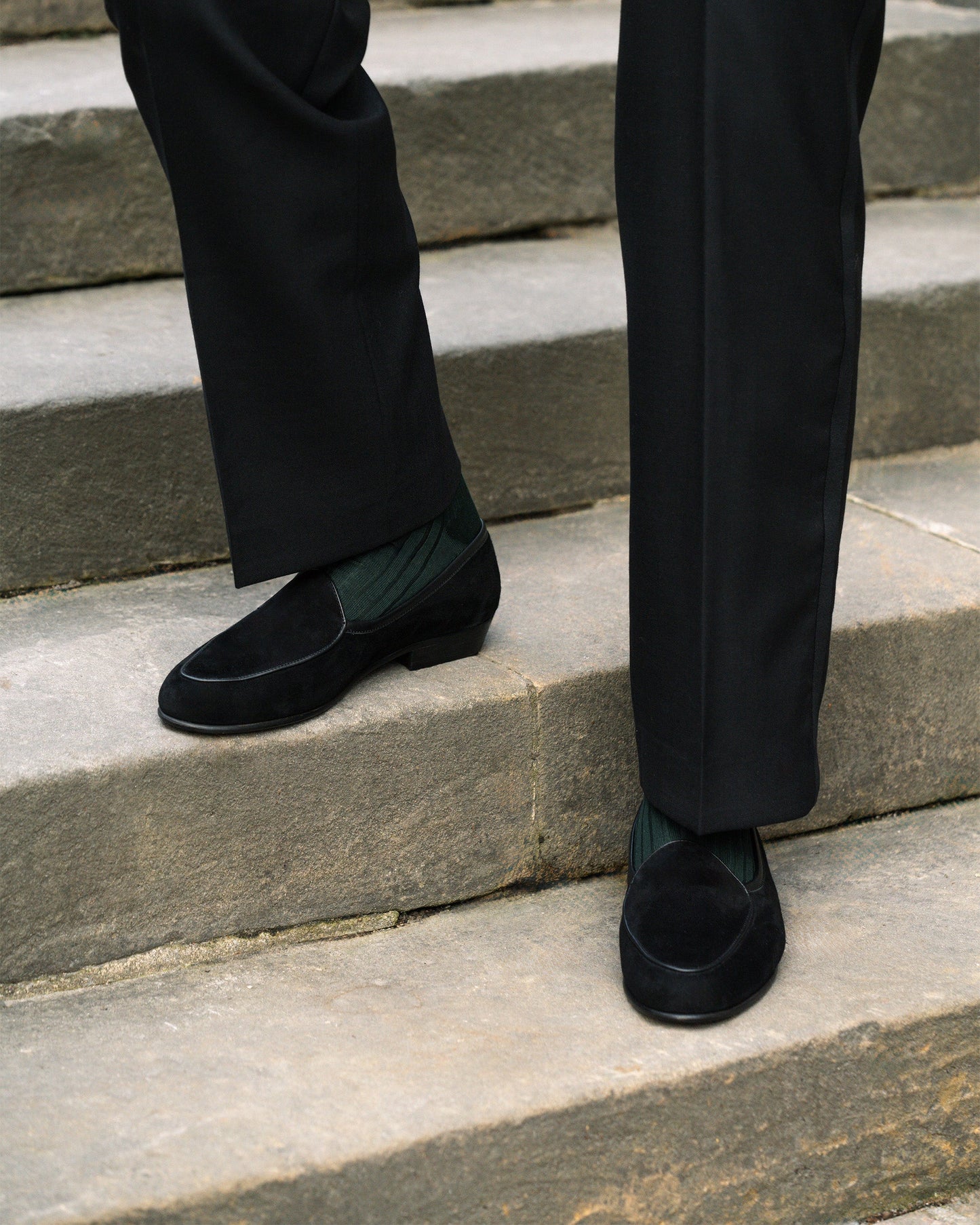 A man wearing Ängby in black suede, styled with tailored black slacks and dark green socks, stands on stone steps, highlighting the loafer’s refined silhouette and versatile elegance for formal or smart casual occasions.