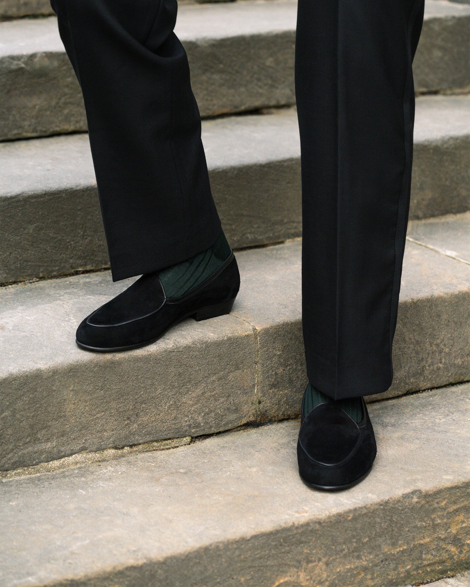 A man wearing Ängby in black suede, styled with tailored black slacks and dark green socks, stands on stone steps, highlighting the loafer’s refined silhouette and versatile elegance for formal or smart casual occasions.