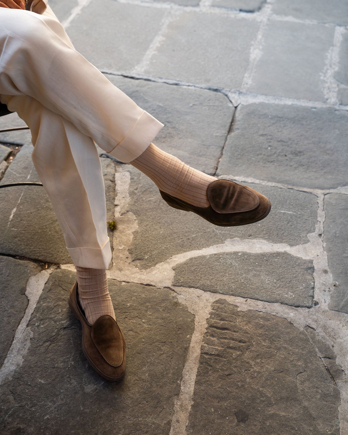 A man sits outdoors wearing Ängby Walker in taupe suede, styled with light-colored trousers and matching socks, highlighting the loafer's minimalist design and versatile shade for refined casual or smart summer looks.