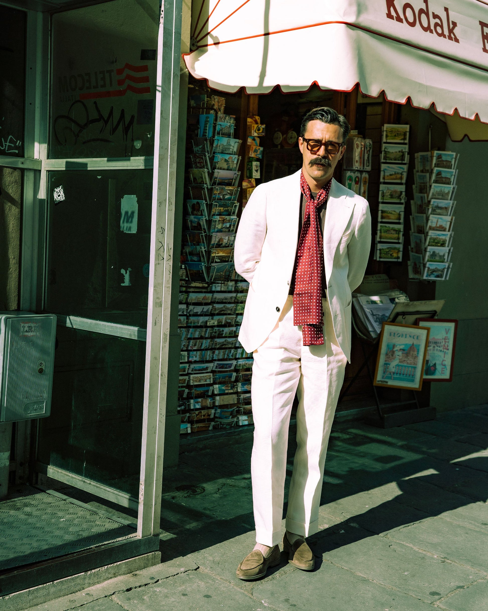 A man stands on a city street wearing Ängby Walker in taupe suede, paired with a white suit and red patterned scarf, showcasing the loafers' versatility and understated style for refined warm-weather occasions.