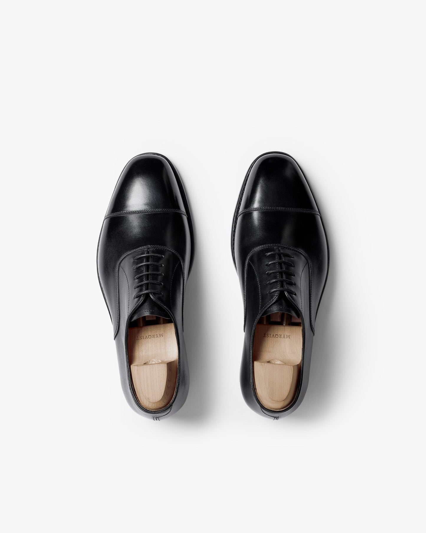 A pair of Äppelviken II in black calf, shown from above, highlights the classic cap-toe Oxford design with closed lacing, polished leather finish, and wooden shoe trees maintaining their elegant shape.