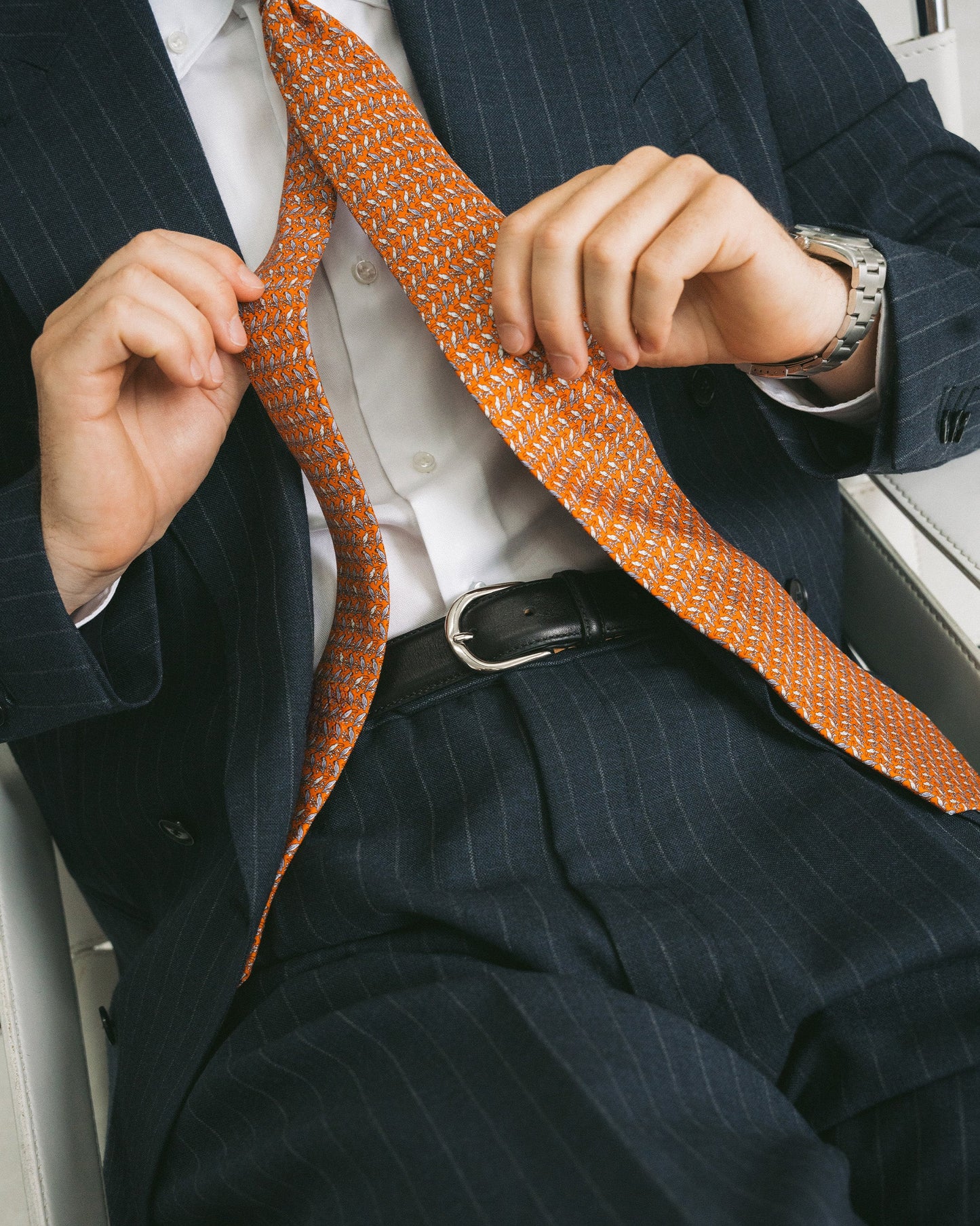 A man wearing a navy pinstripe suit and orange patterned tie is styled with a black leather belt featuring a polished silver buckle, creating a refined look suitable for business or formal occasions.
