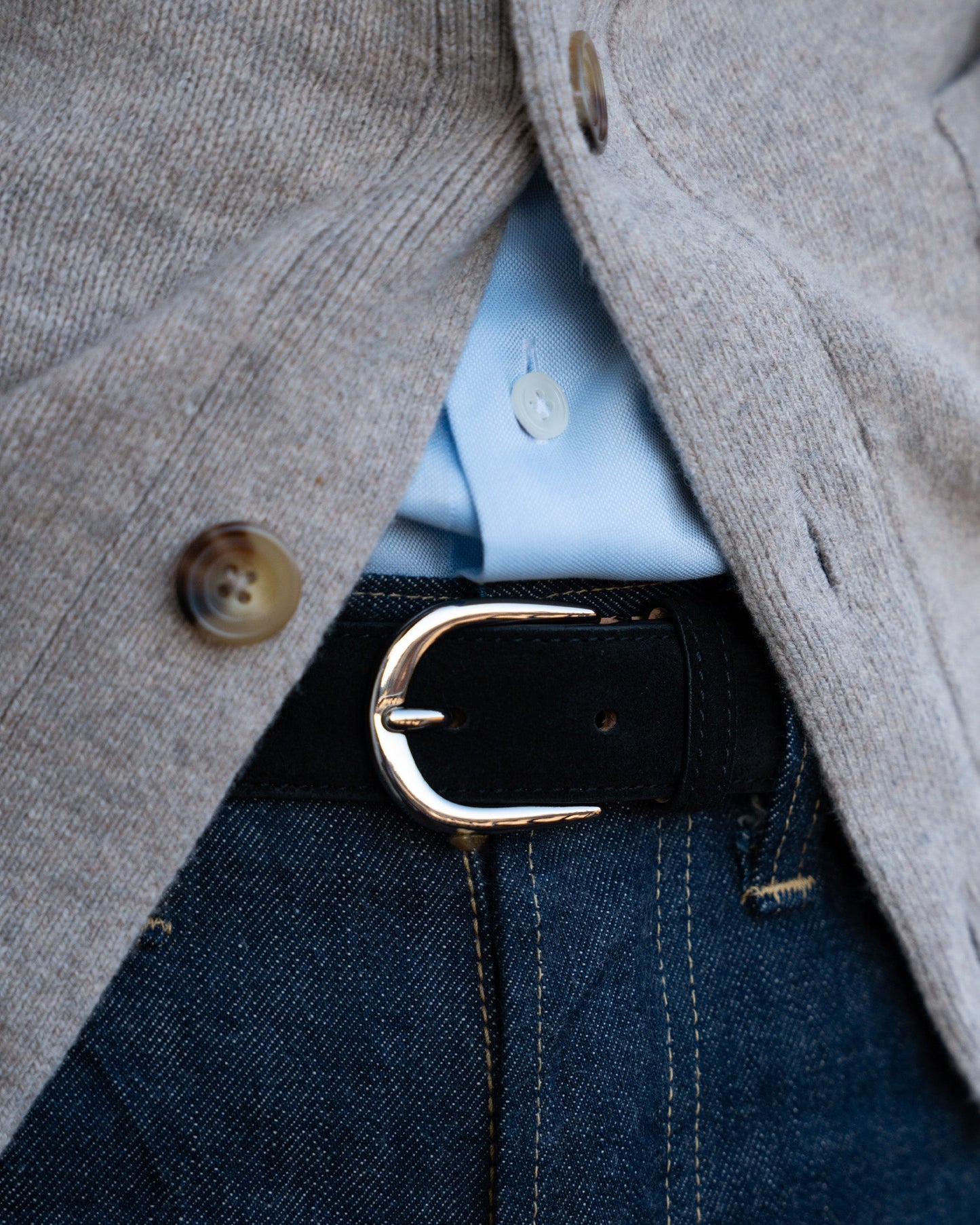 Black suede belt with a round silver buckle, styled with blue denim jeans, a light blue shirt, and a beige cardigan for a refined yet relaxed look.