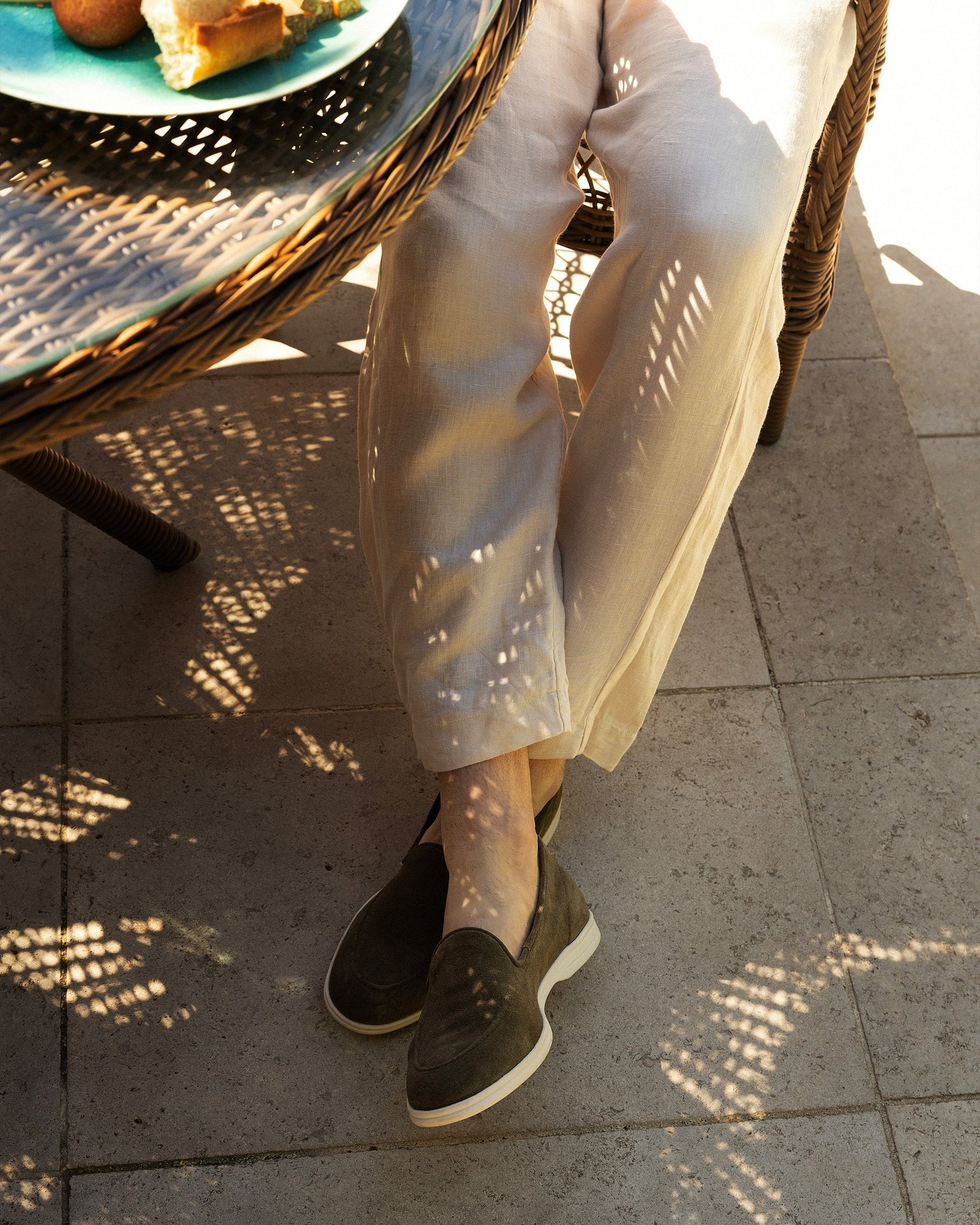 Brännö II in moss green suede worn with light linen trousers, shown in a relaxed outdoor setting with sunlight filtering through a wicker table, highlighting the loafer's soft suede texture and white rubber sole.