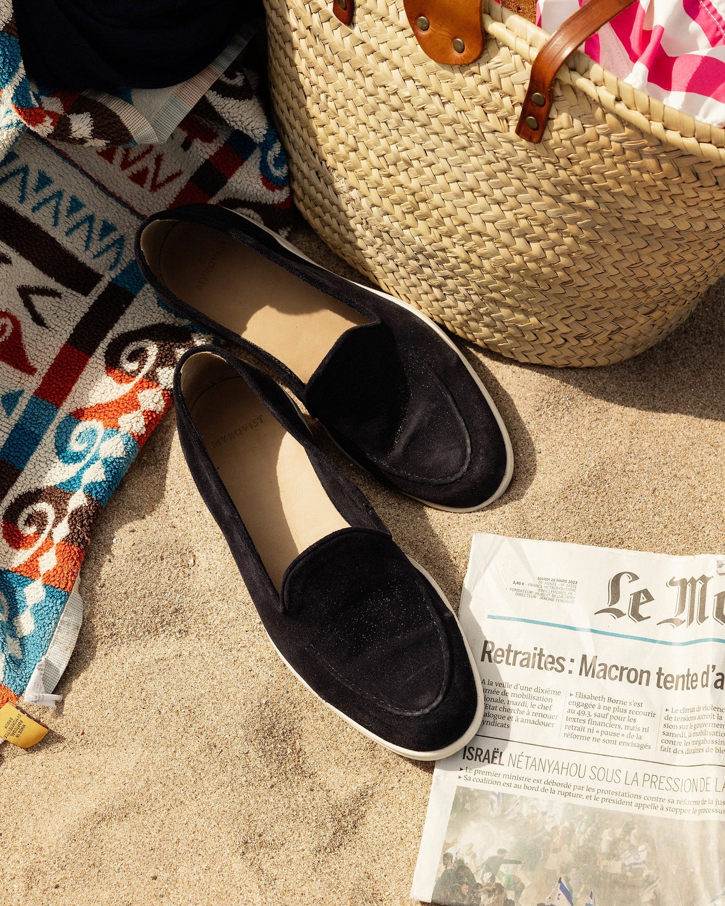 Brännö II in navy suede loafers rest on sandy beach beside a woven tote, patterned towel, and newspaper, showcasing their deep navy suede uppers and white rubber soles in a relaxed summer setting.