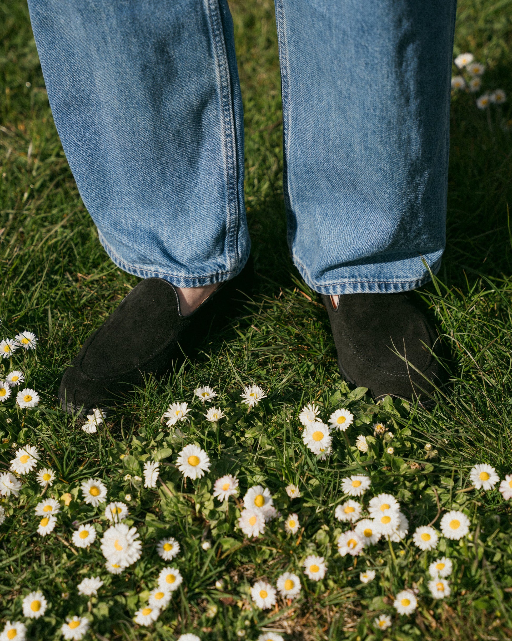 Brännö II in black suede worn with relaxed denim, shown standing on grass dotted with white daisies for a casual spring or summer look.