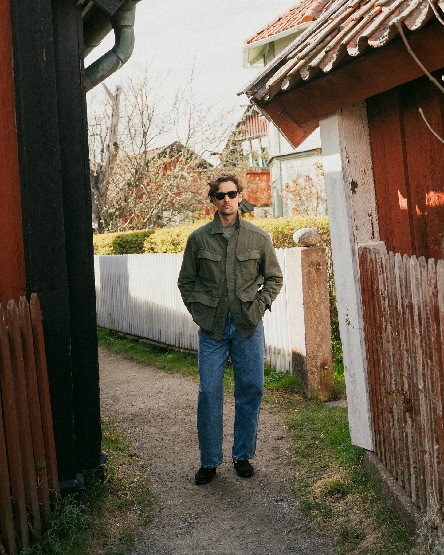 A man wears Brännö II in black suede with relaxed jeans and an olive jacket, standing on a narrow path between rustic wooden houses in a casual outdoor setting.