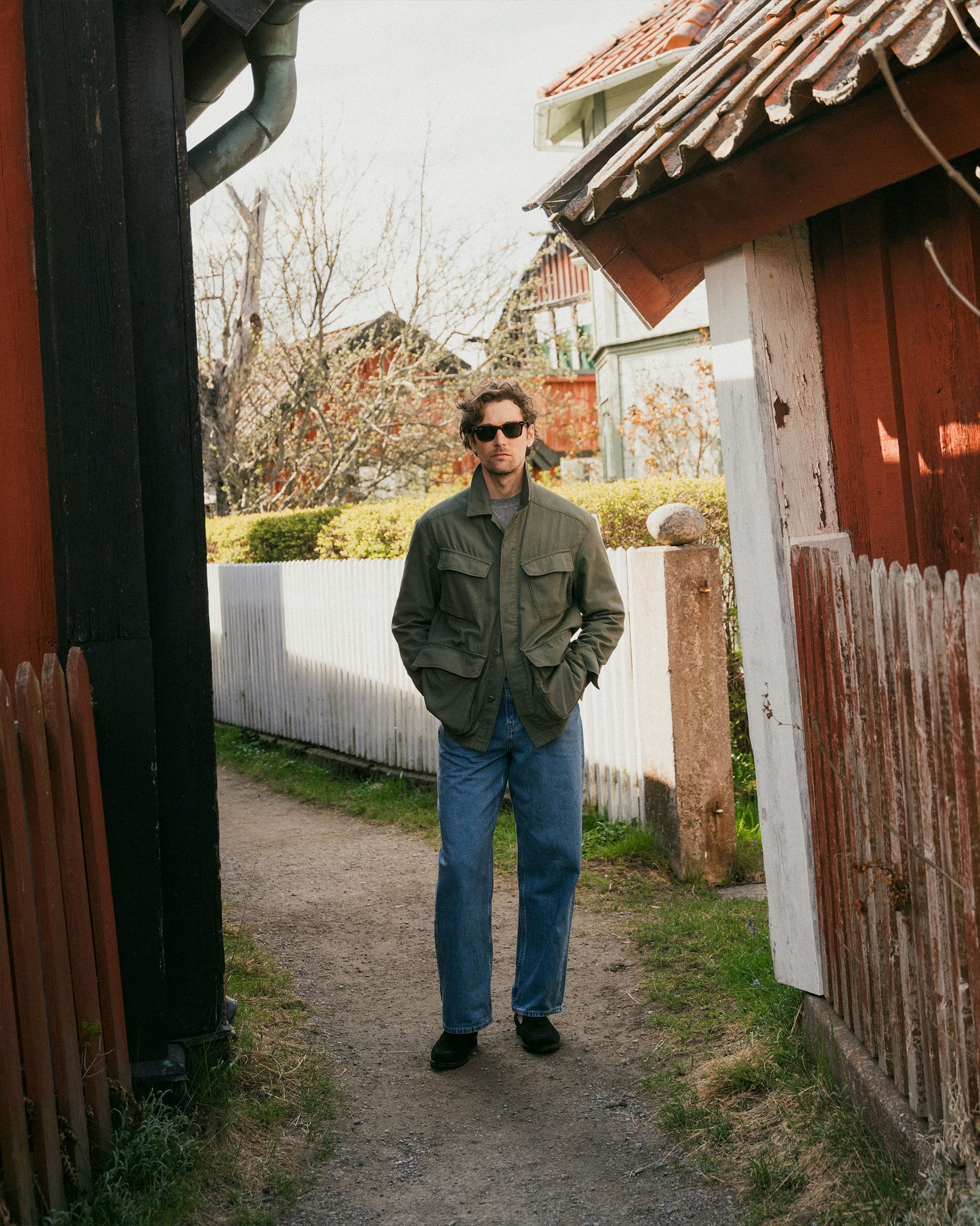 A man wears Brännö II in black suede with relaxed jeans and an olive jacket, standing on a narrow path between rustic wooden houses in a casual outdoor setting.