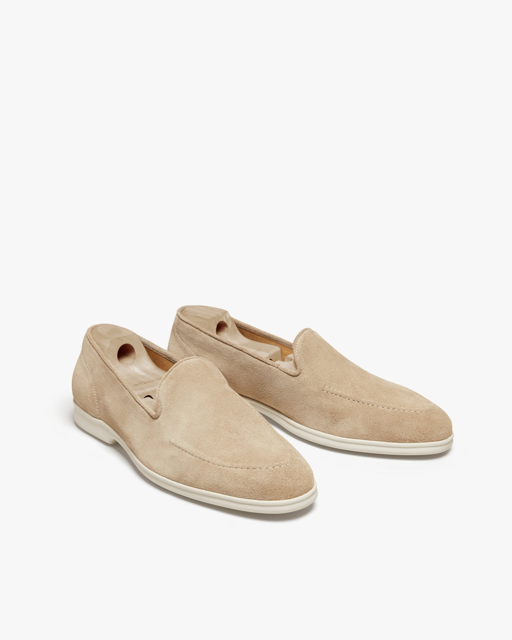 Brännö II in sand suede showcases a pair of minimalist men's loafers with a soft beige suede upper, clean stitching, and white rubber soles, presented with wooden shoe trees for shape and care.