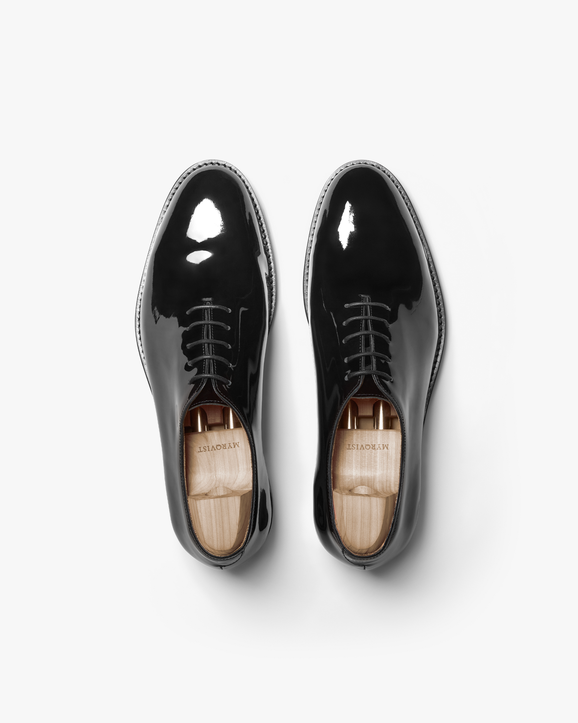 A pair of Drottningholm in black patent leather shown from above, highlighting the whole-cut Oxford construction, glossy mirror-like finish, almond-shaped toes, and wooden shoe trees inside each shoe.