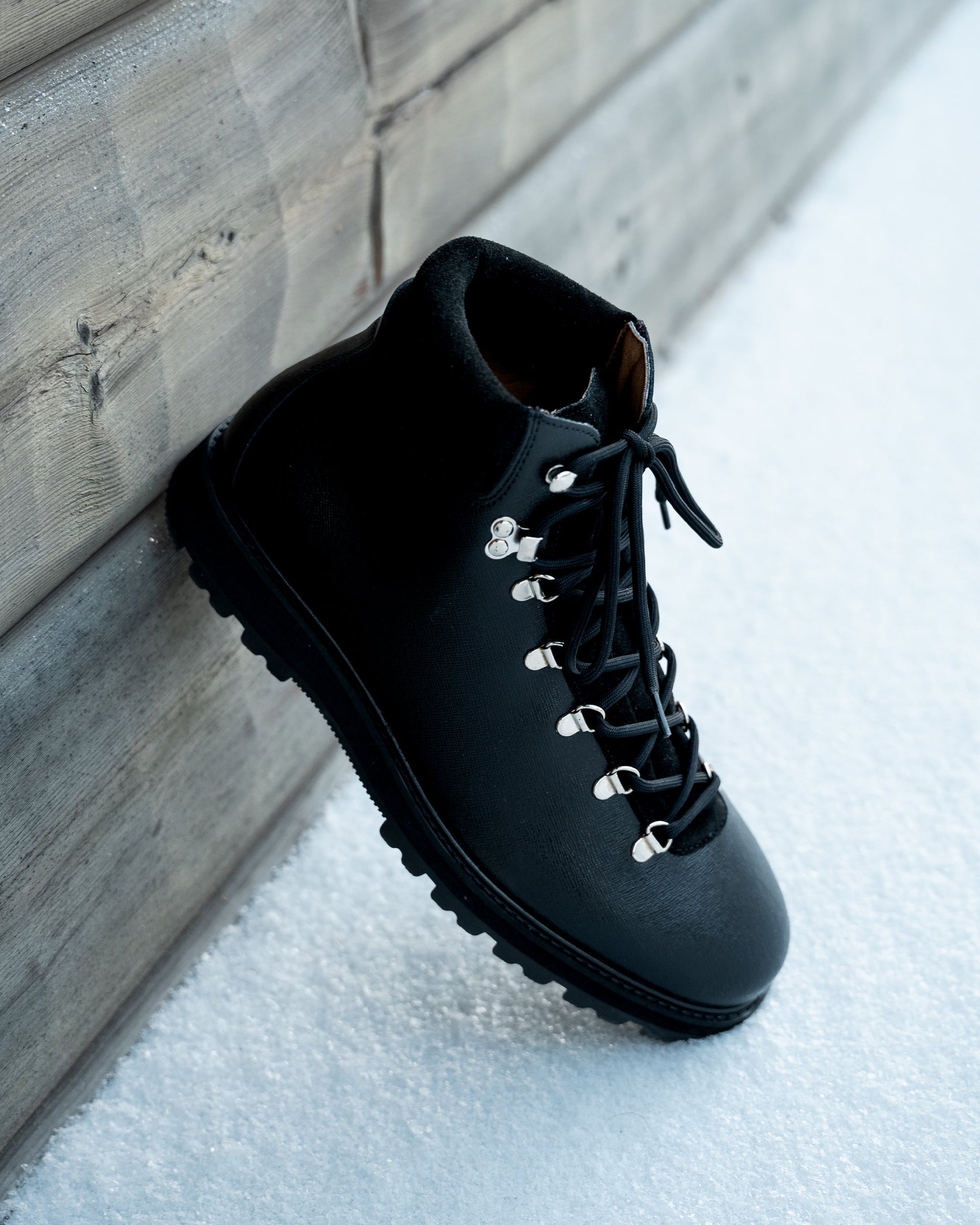 Duved in black grain calf displayed outdoors on snow, showcasing its textured leather, silver-toned hardware, and robust hiking sole designed for winter conditions.