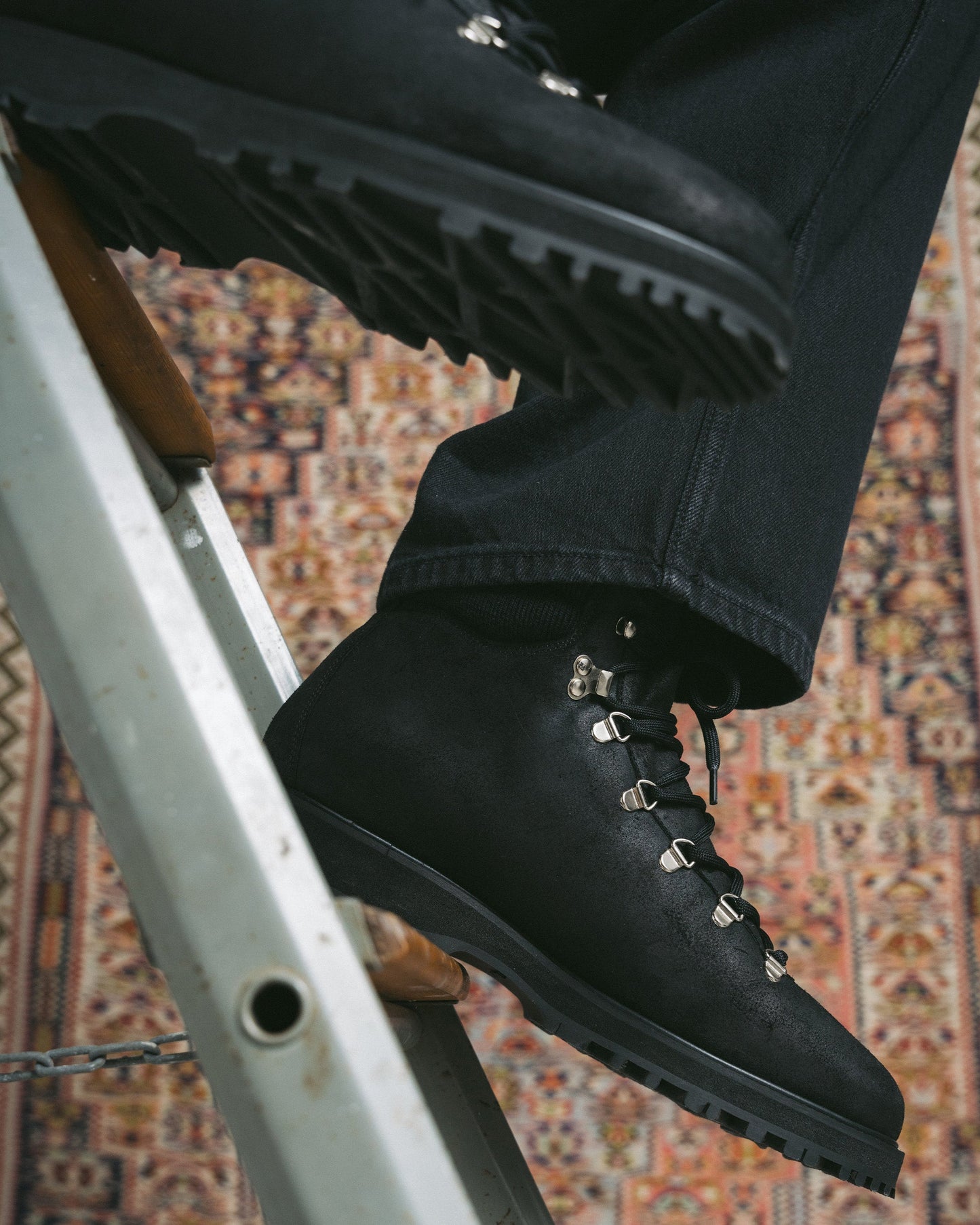 Duved in black waxed suede is worn with dark denim, highlighting its water-repellent suede, metal eyelets, and rugged hiking sole, shown against a patterned rug for a versatile, urban-inspired look.