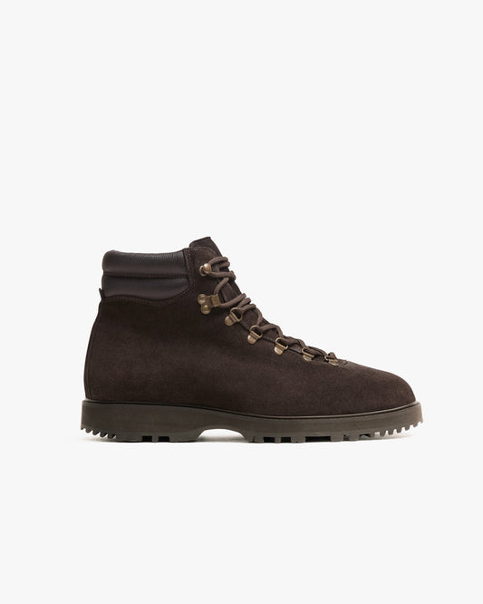 Duved in dark brown suede features a rugged hiking boot silhouette with brass-toned hardware, a padded leather collar, and a sturdy lug sole, highlighting its blend of refined style and outdoor functionality.