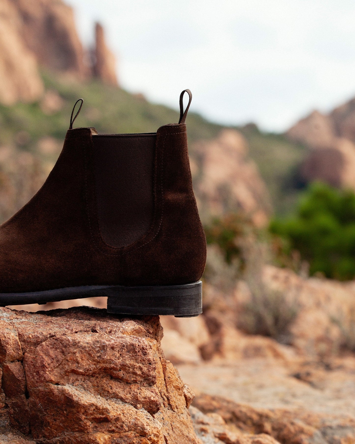 Elfvik Chelsea boot in dark brown suede is shown resting on a rocky surface, highlighting its water-repellent suede, elastic side panel, and sturdy sole designed for versatile outdoor wear.