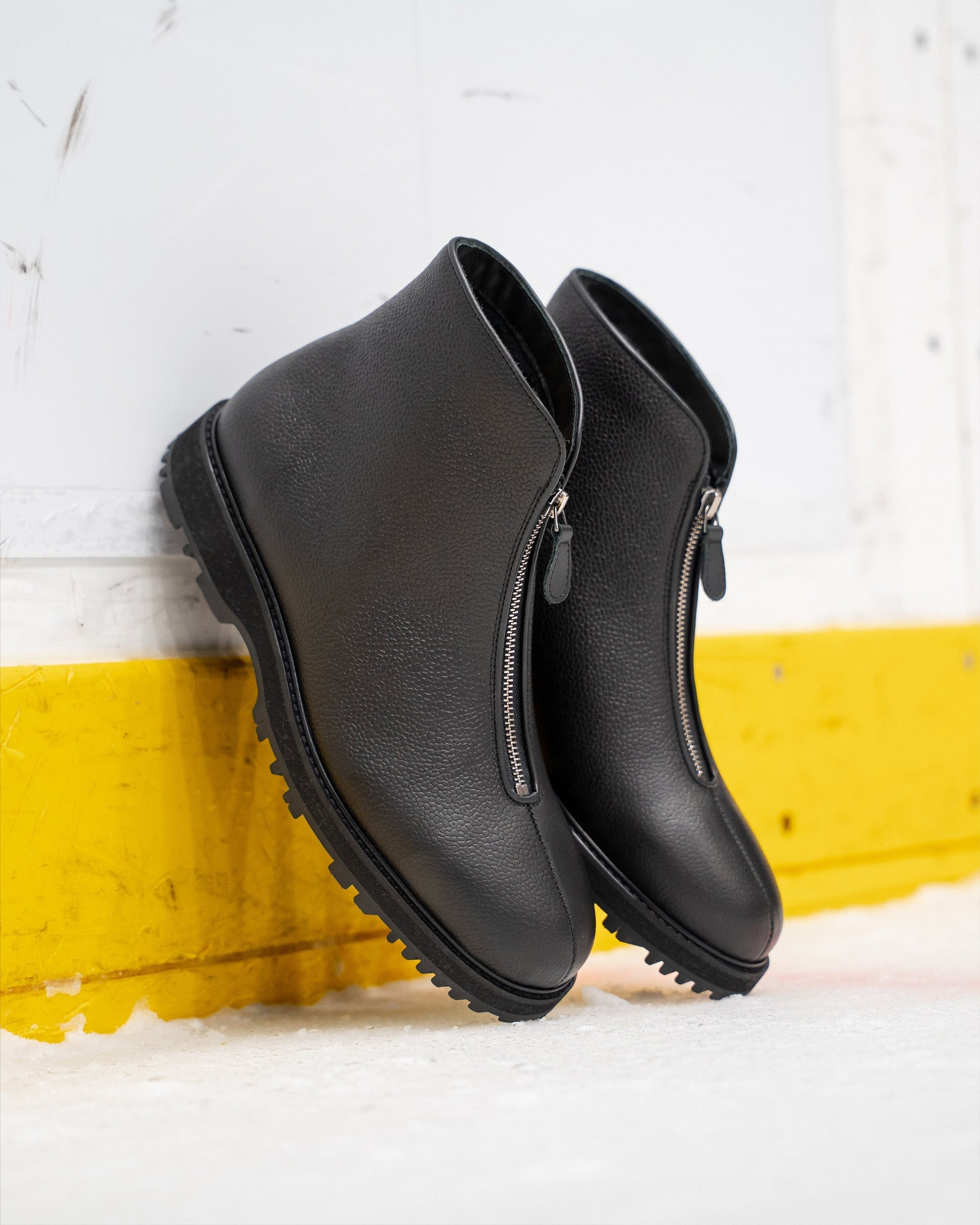 Funäs in black country calf features a pair of textured leather boots with central zippers and rugged Explorer soles, shown leaning against a rink barrier for a modern winter look.