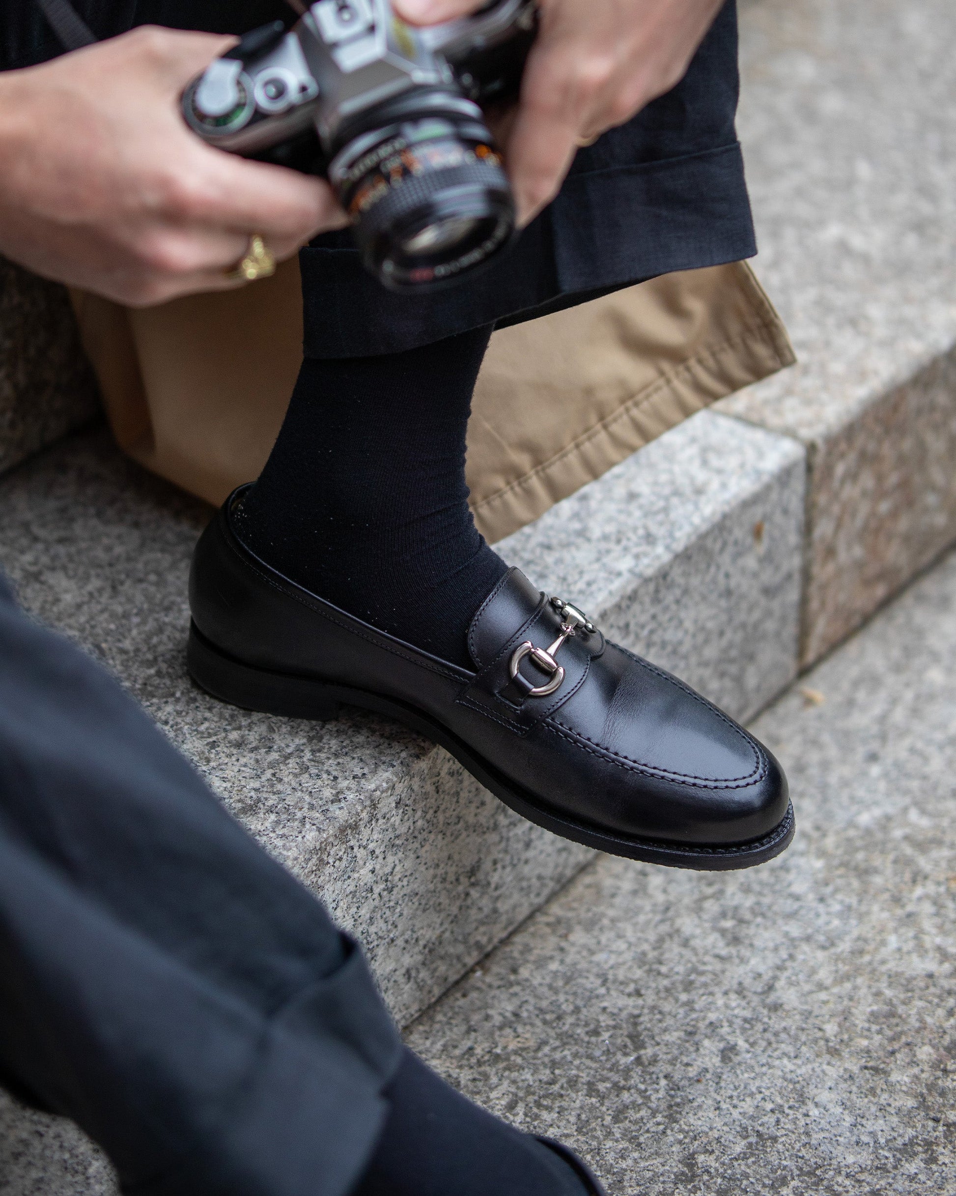 Karlberg in black calf leather worn with dark trousers and black socks, featuring a polished finish and silver-toned horsebit detail, styled for a casual city setting with a camera in hand.