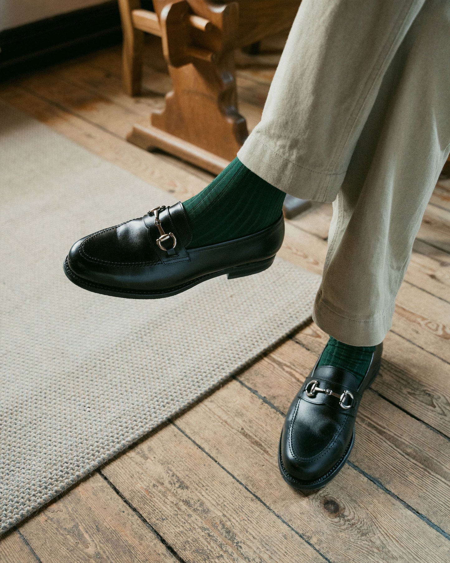 Karlberg in black calf leather shown on foot with tailored light trousers and green socks, highlighting the loafer’s polished finish, rounded toe, and silver-toned horsebit detail in a relaxed indoor setting.