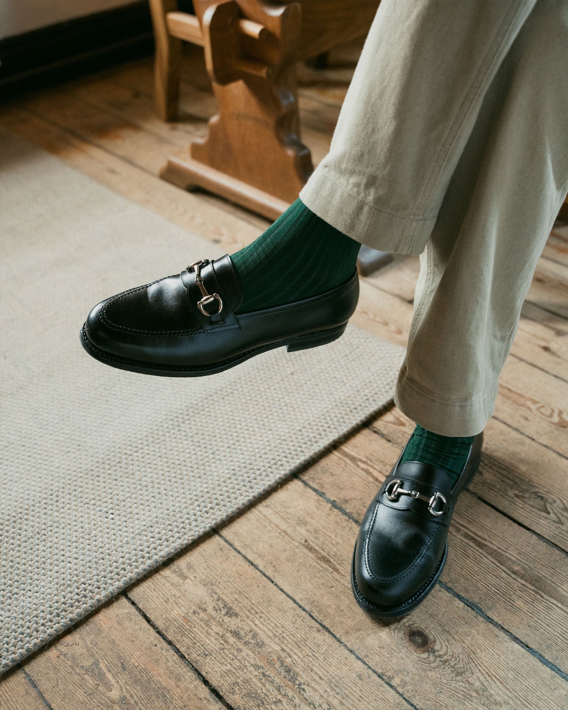Karlberg in black calf leather shown on foot with tailored light trousers and green socks, highlighting the loafer’s polished finish, rounded toe, and silver-toned horsebit detail in a relaxed indoor setting.