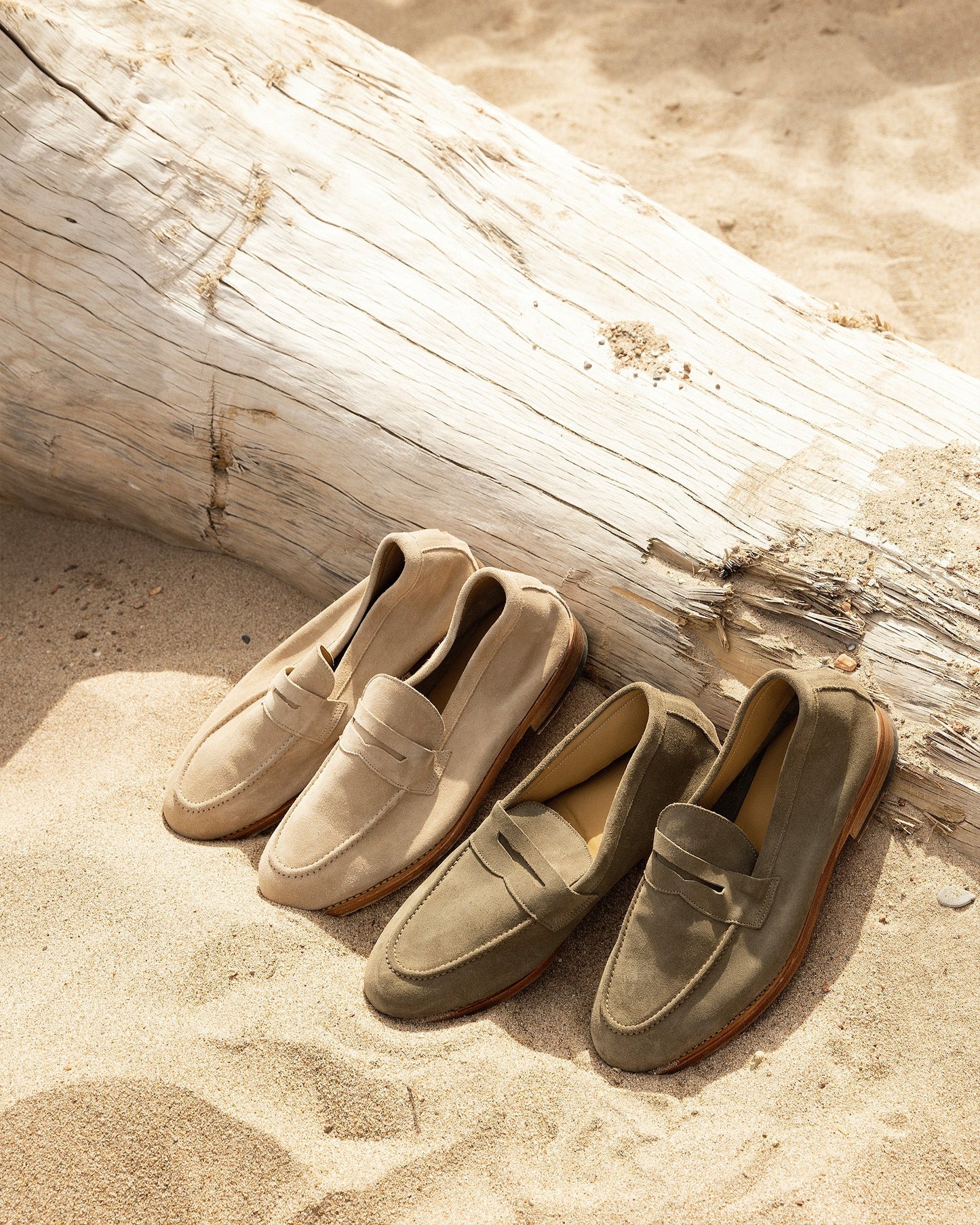 Lysekil II in green suede is displayed alongside a sand-colored pair, both resting on sunlit sand beside a weathered driftwood log, highlighting the loafers’ soft suede texture and relaxed, coastal-inspired style.