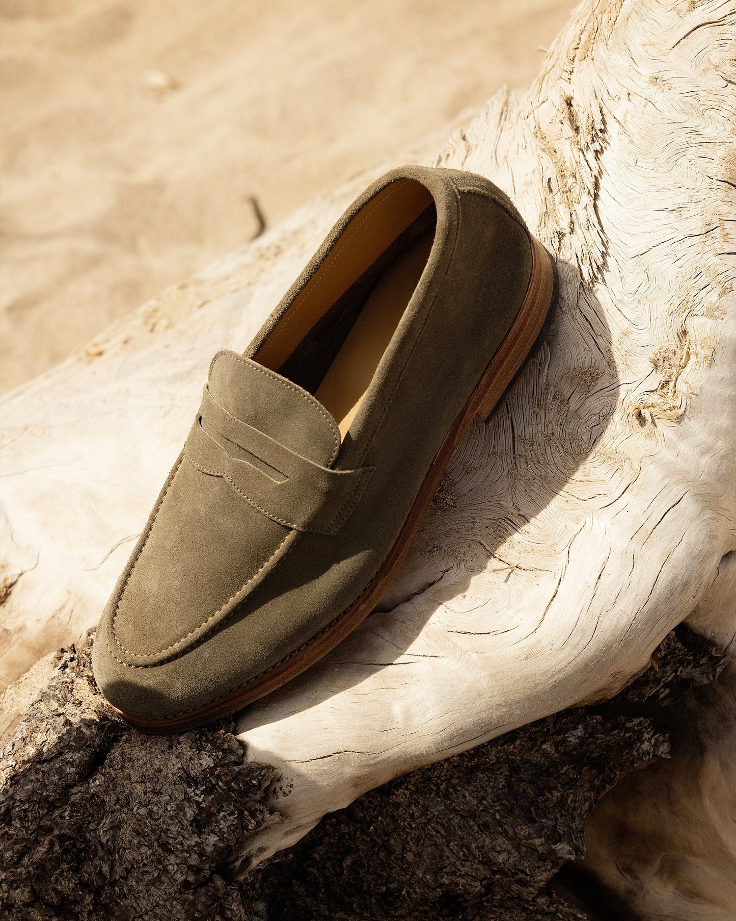 Lysekil II in green suede is shown resting on sun-bleached driftwood, highlighting the loafer’s soft suede texture, penny strap, and relaxed Scandinavian-inspired style ideal for warm weather.