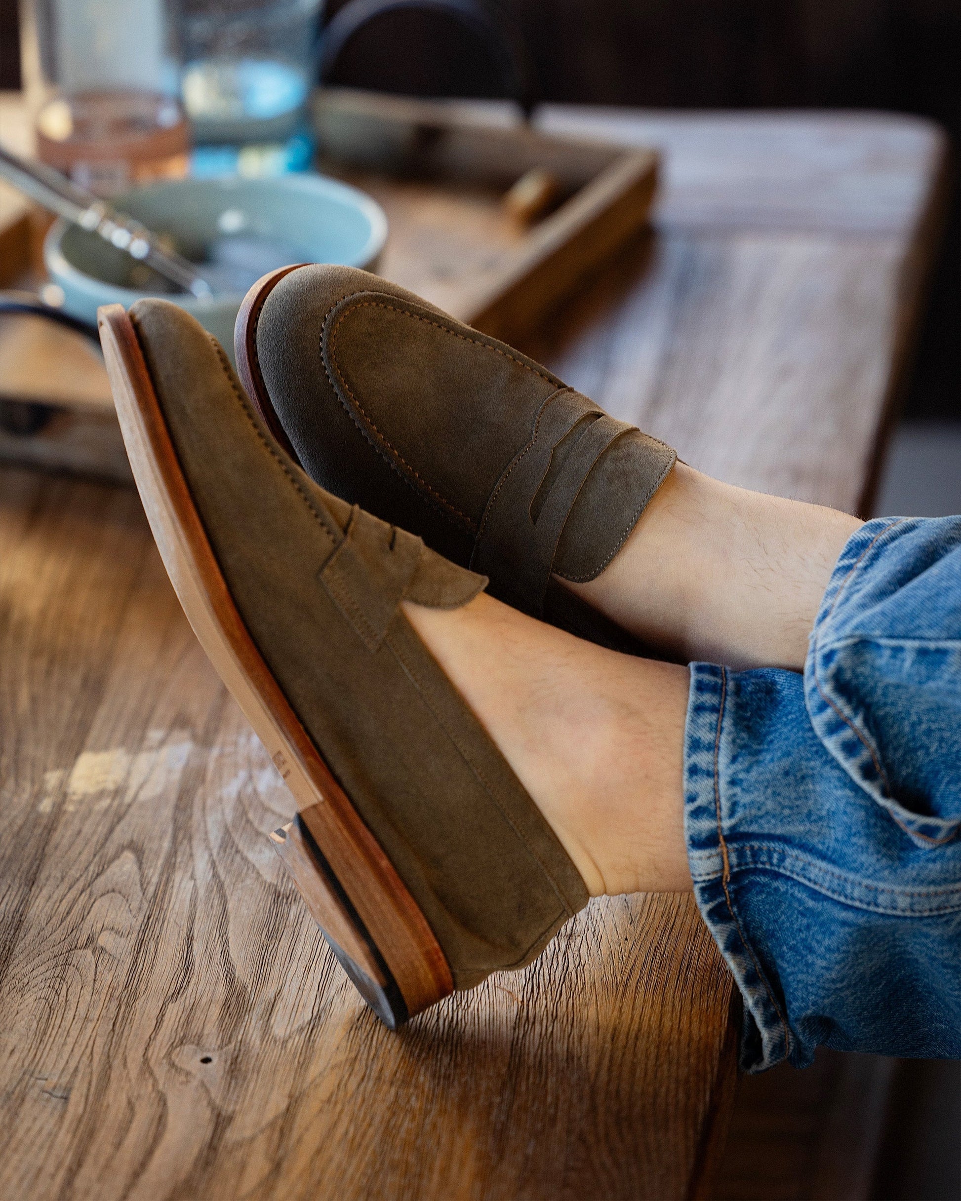 A man relaxes indoors with his feet up, wearing Lysekil II in green suede styled with casual denim, showcasing the loafer’s soft suede finish, penny strap, and versatile silhouette.
