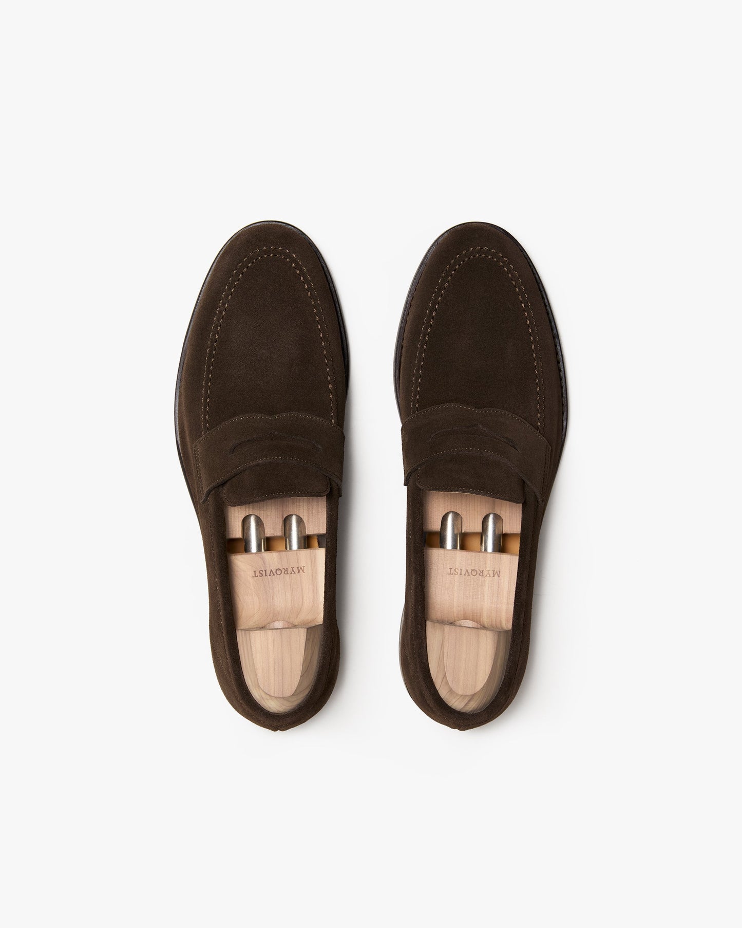 A pair of Lysekil II penny loafers in dark brown suede, shown from above with wooden shoe trees inserted, highlighting the apron stitching, rounded toe, and soft suede texture.