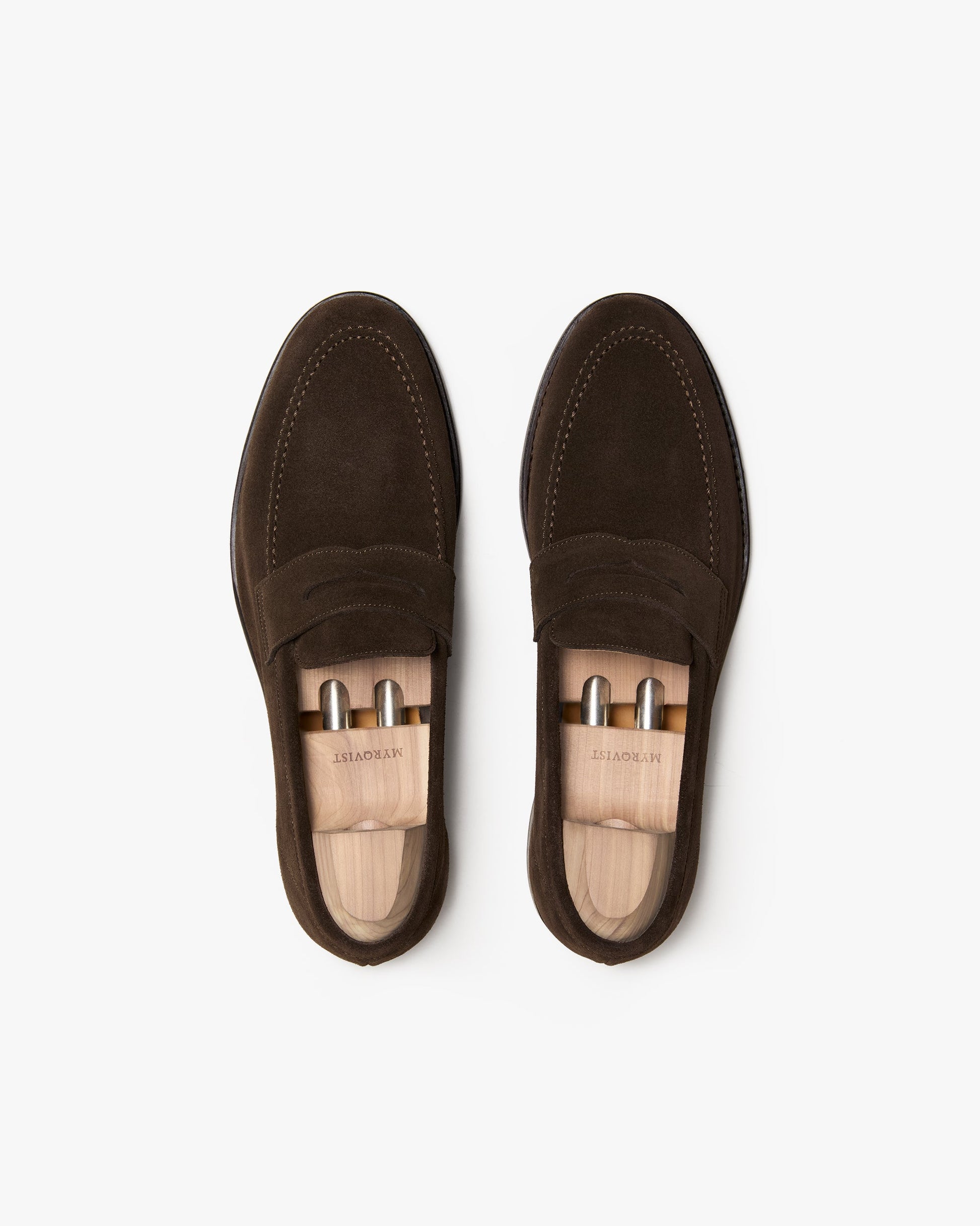 A pair of Lysekil II penny loafers in dark brown suede, shown from above with wooden shoe trees inserted, highlighting the apron stitching, rounded toe, and soft suede texture.