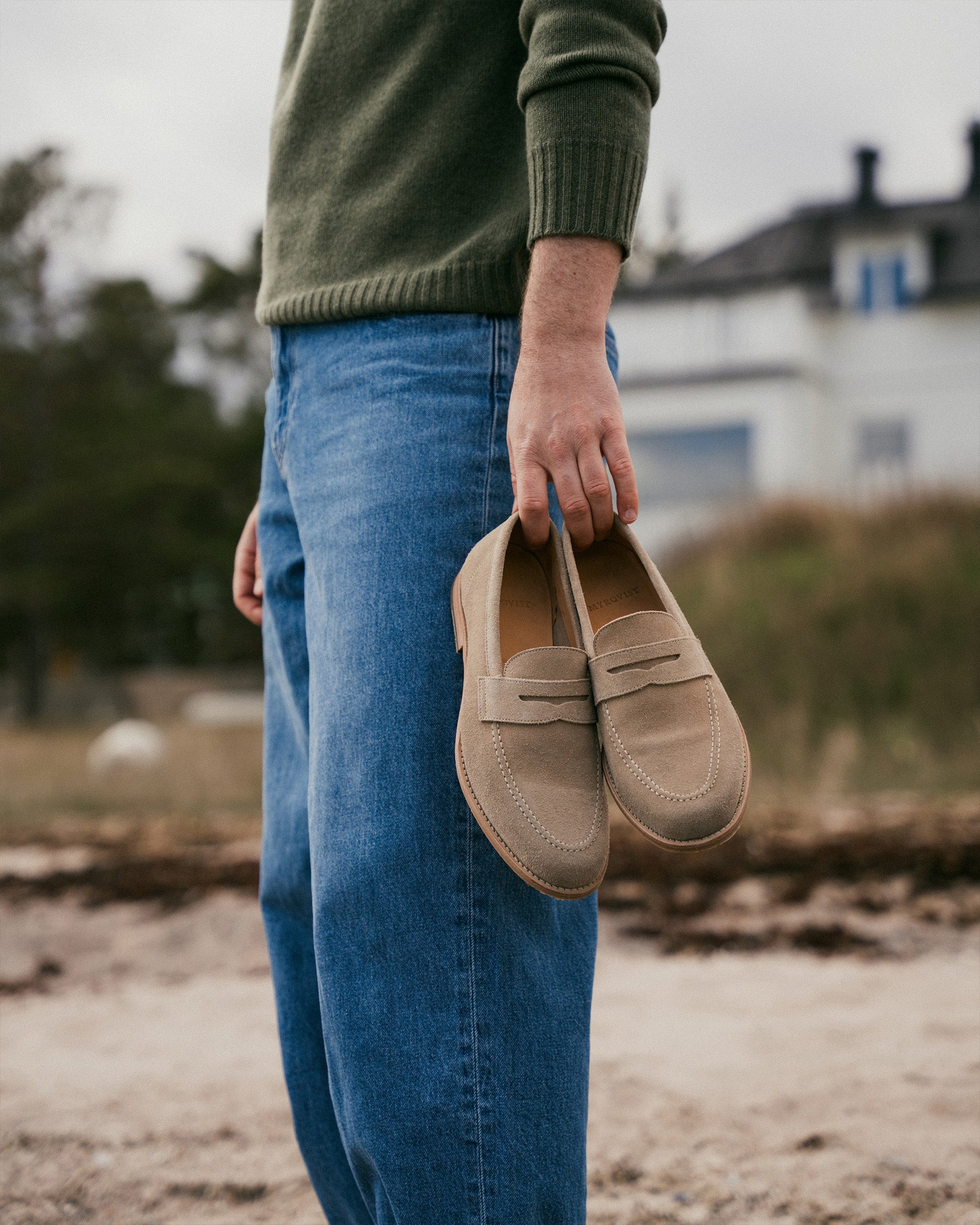 A man stands outdoors in casual jeans and a green sweater, holding a pair of Lysekil II in sand suede penny loafers by his side, highlighting the soft suede texture and classic design in a relaxed, natural setting.