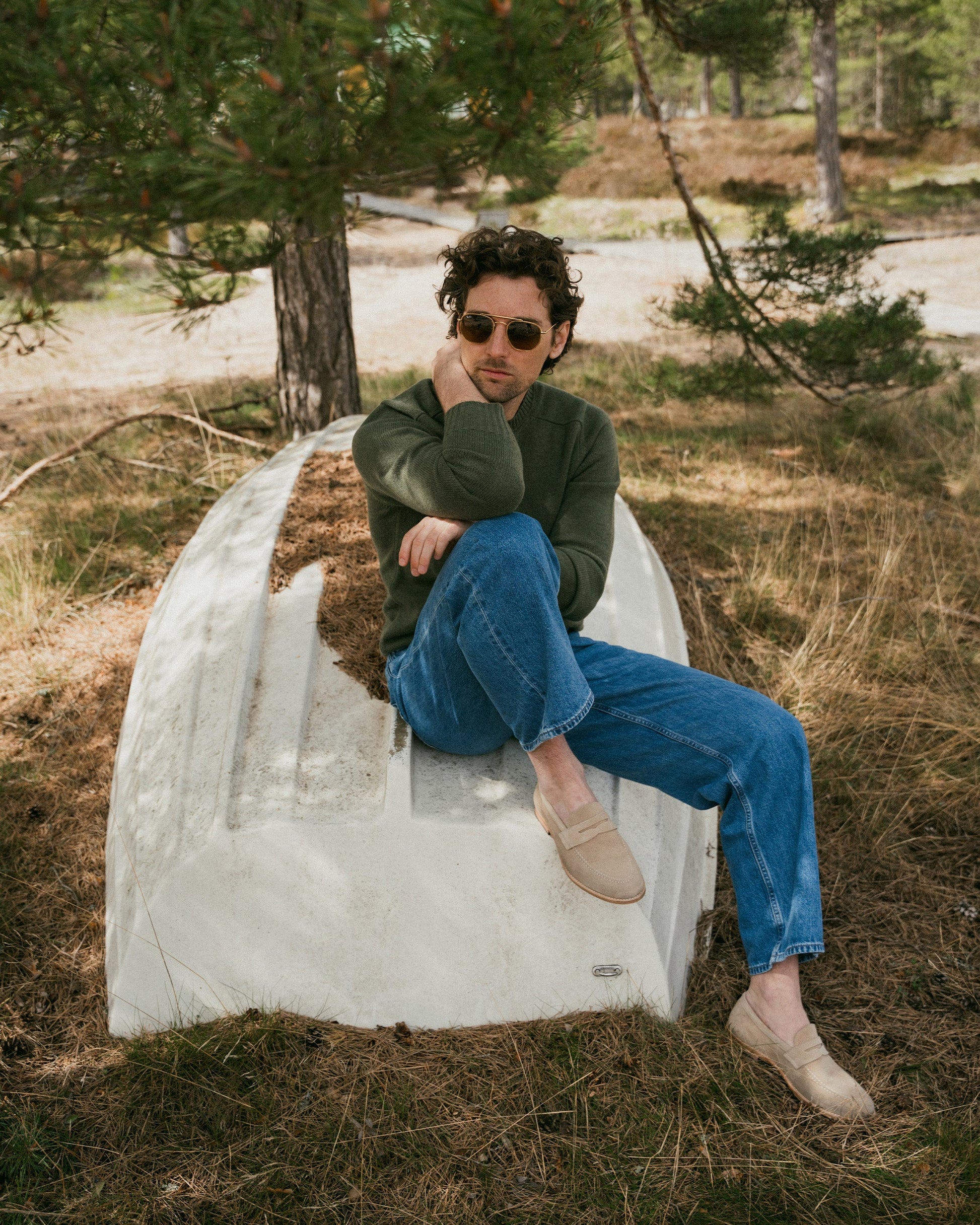 A man sits outdoors on an overturned boat in a wooded setting, wearing Lysekil II in sand suede penny loafers paired with blue jeans and a green sweater, showcasing the relaxed and versatile style of these suede loafers for casual occasions.