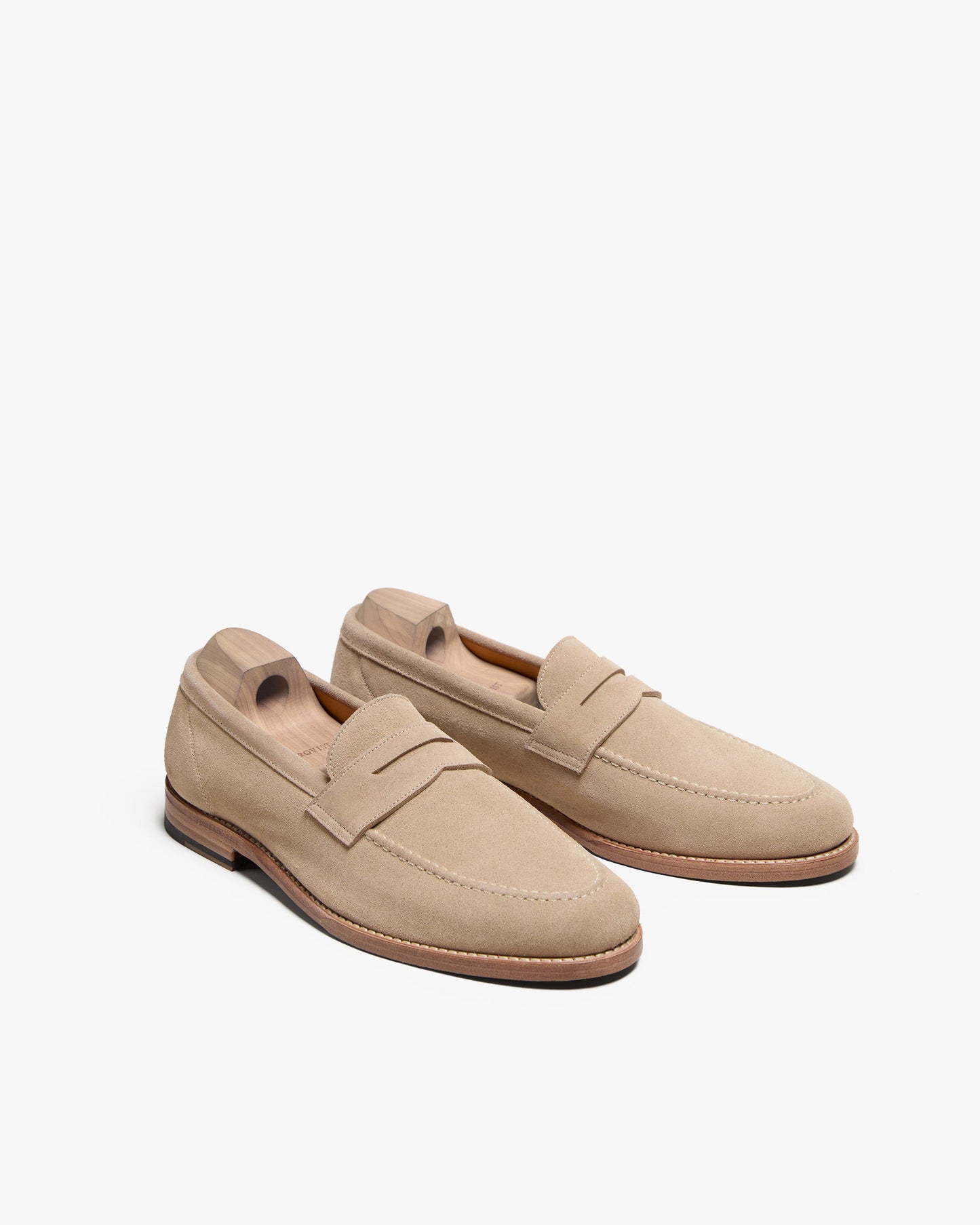 Lysekil II in sand suede penny loafers displayed at a slight angle, featuring soft suede uppers, classic penny strap, apron stitching, and wooden shoe trees to maintain their elegant shape.