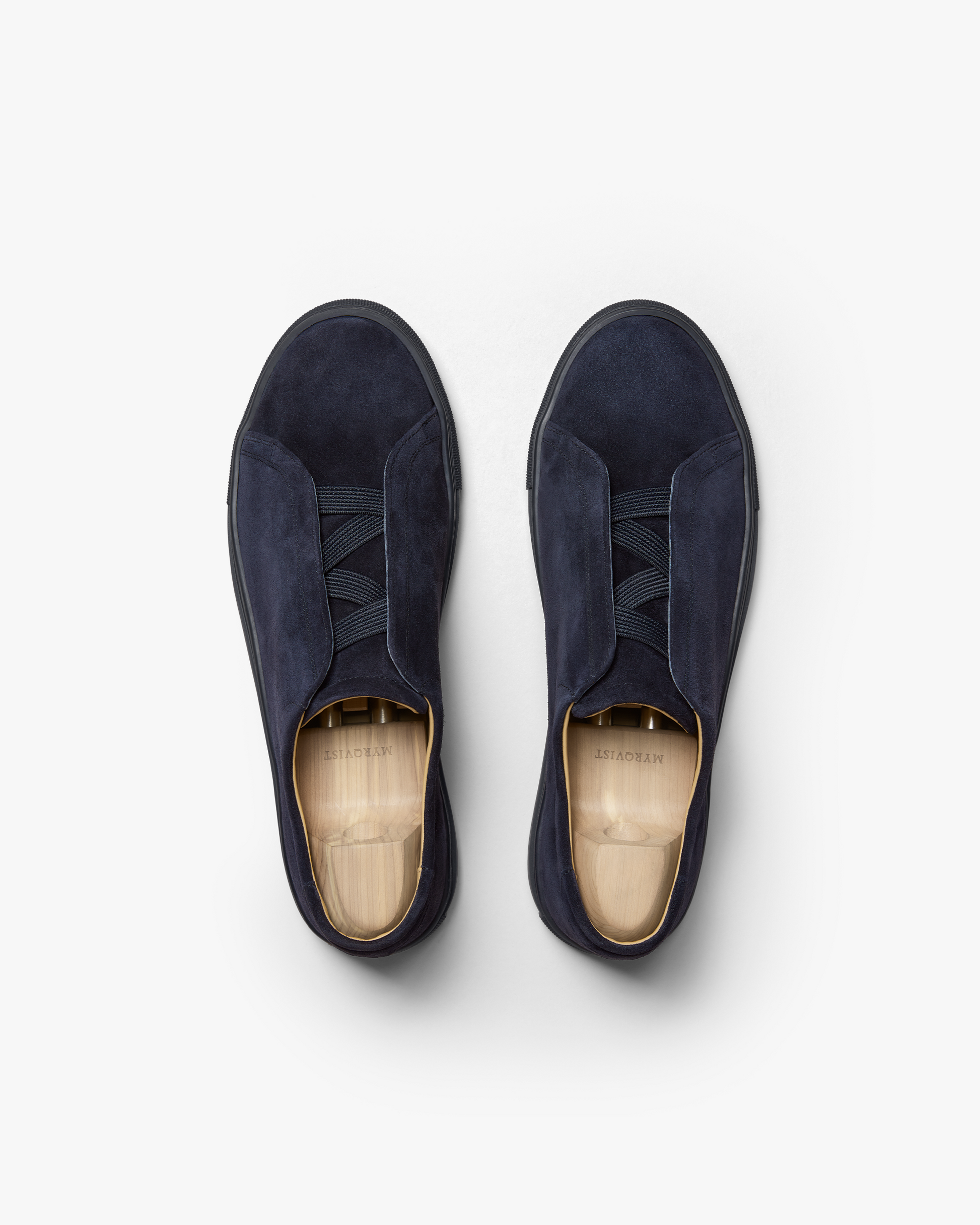 Pair of Alnö Monochrome II slip-on sneakers in midnight blue suede shown from above, featuring seamless uppers, tonal elastic across the instep, and wooden shoe trees inside each shoe.