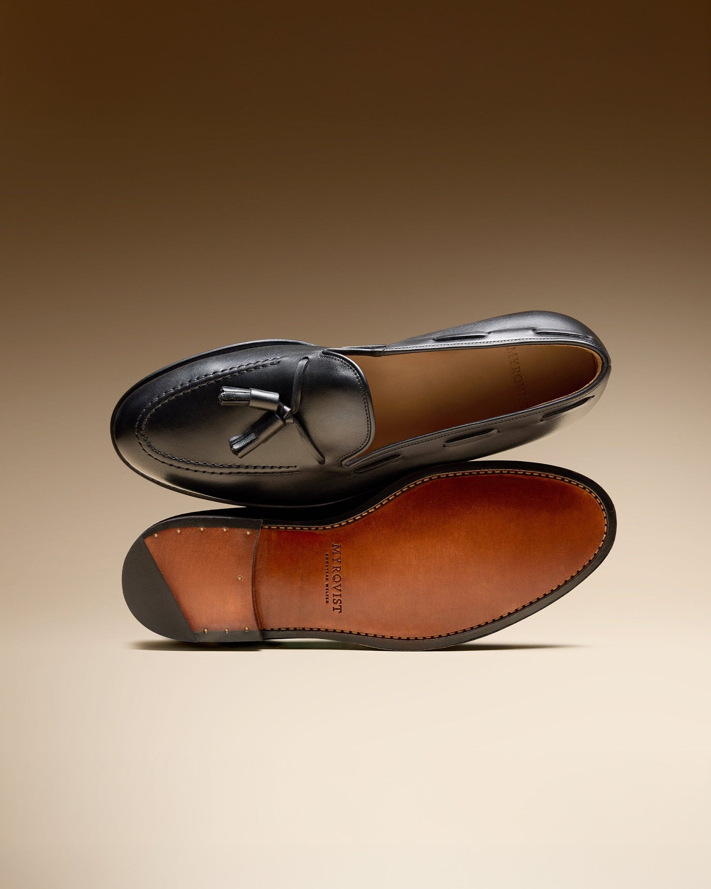 Mölle II in black calf displayed with one shoe showing the polished tassel loafer upper and the other highlighting the full leather sole with Goodyear welted construction and Myrqvist branding, set against a warm gradient background.