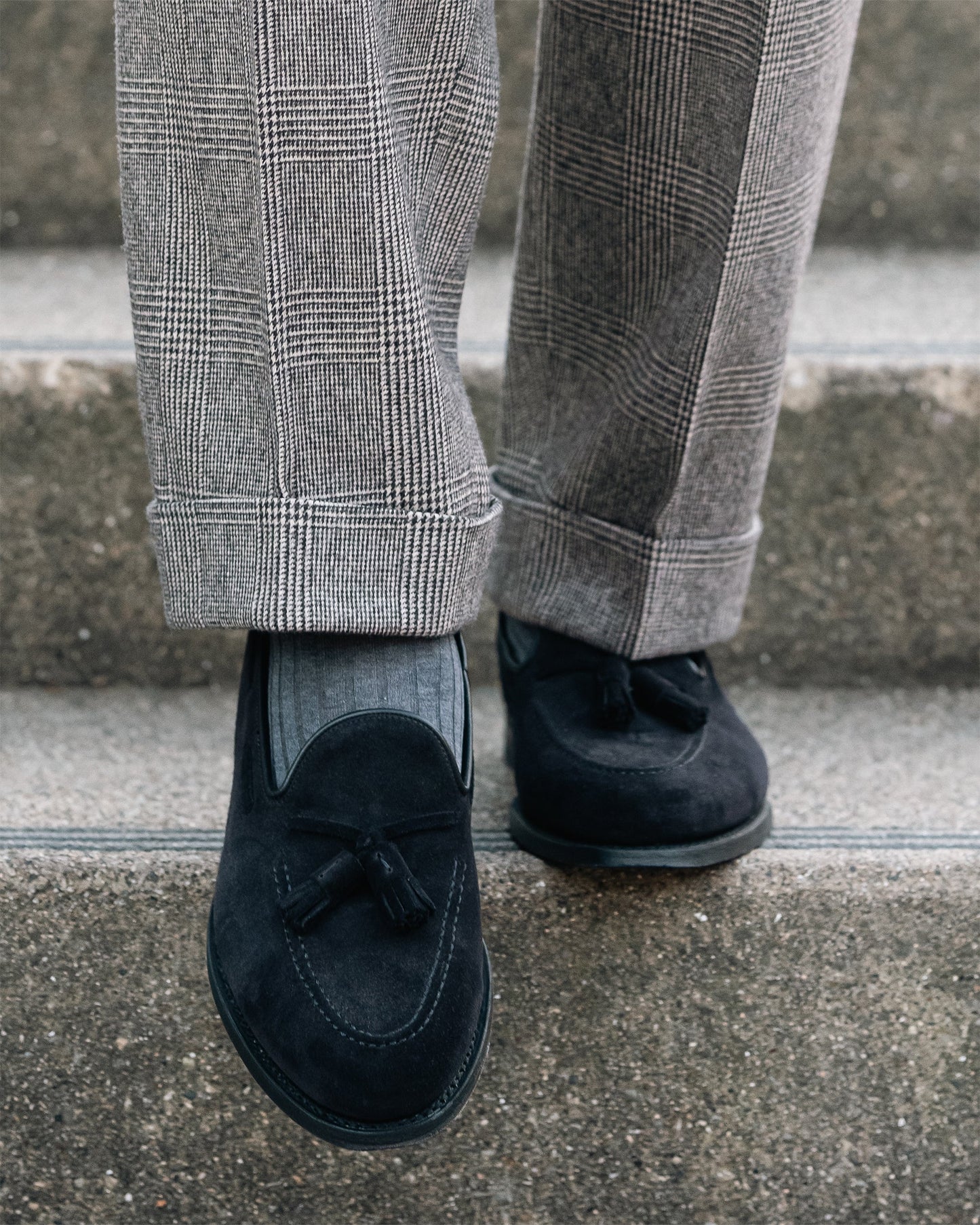 Mölle II in black suede tassel loafers styled with gray plaid slacks, highlighting the soft suede texture, apron front, and refined stitching in an urban setting.