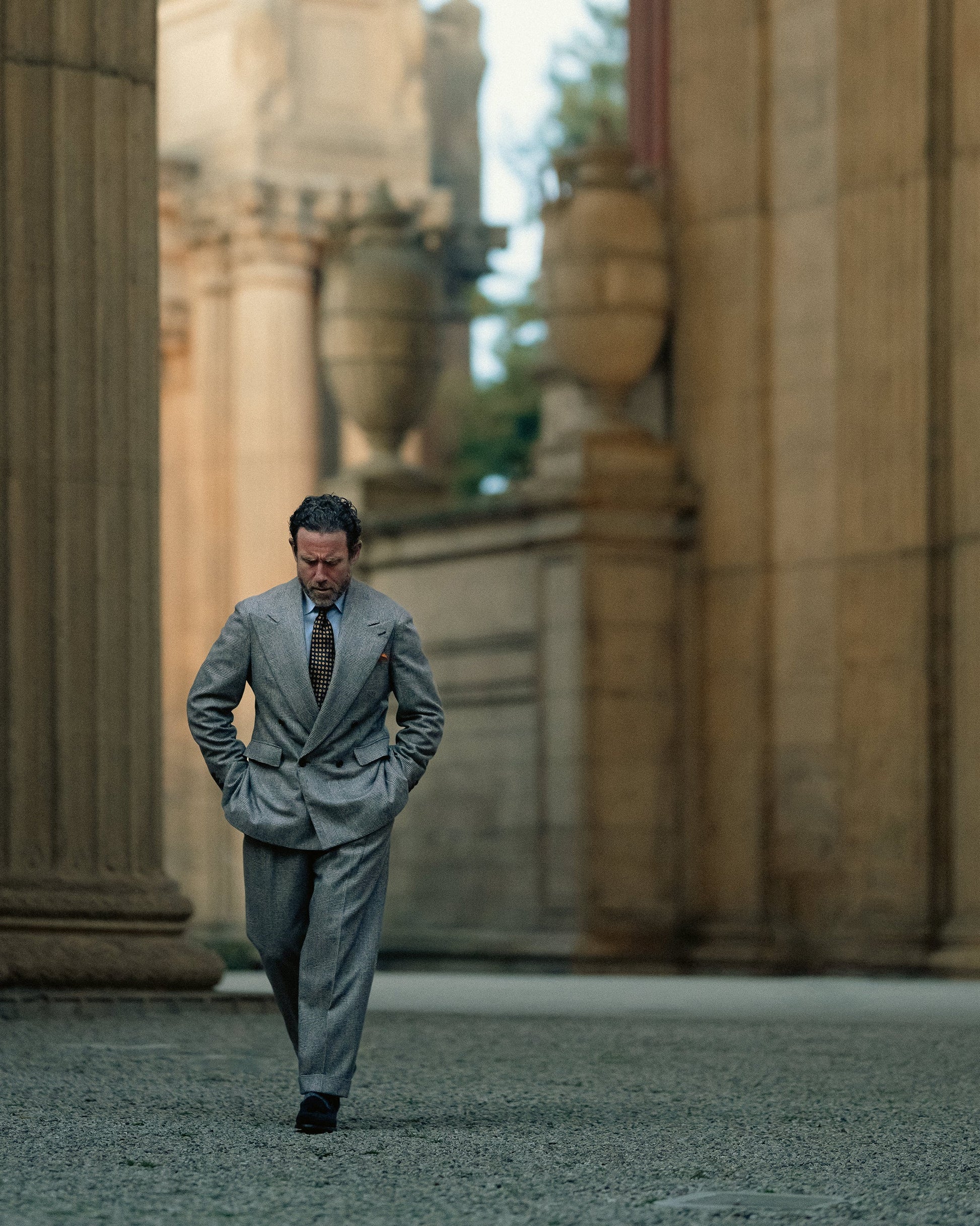 A man wearing Mölle II in black suede walks confidently through a grand architectural setting, pairing the black suede tassel loafers with a tailored gray suit for an elegant, formal look.